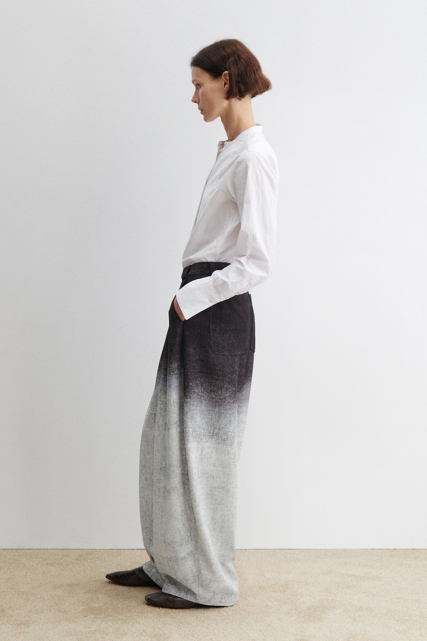 Full length side view of a woman standing wearing spring fashion in the Rachel Comey black Dias pant featuring a high waist and wide-leg silhouette, cut from softly textured fabric with a natural drape and ombre effect. Front pleats add volume and ease, creating a relaxed fit through the leg at Alas Eius.