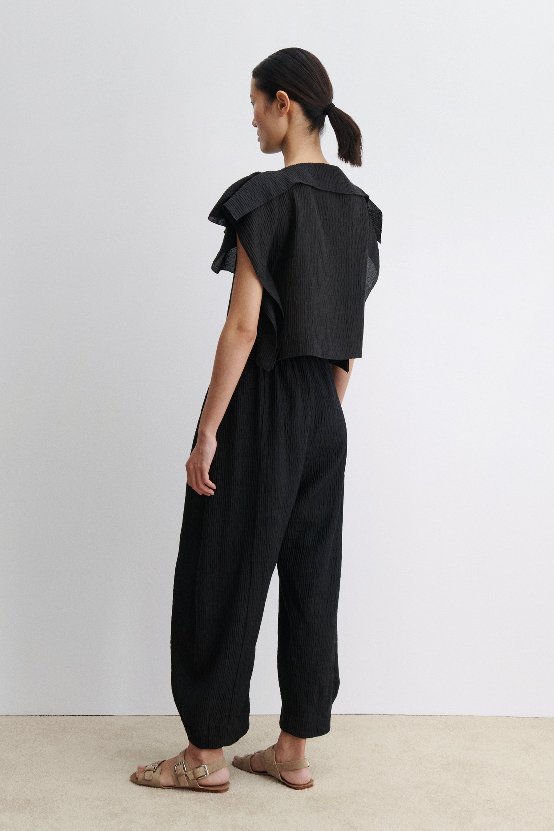 3/4 back view of a woman standing wearing contemporary spring and summer womenswear fashion in a coordinating set by Rachel Comey. The airy Black Rivera Top features softly crinkled fabric, cascading flutter sleeves, and a relaxed, boxy silhouette. Perfectly paired with coordinating Ashbolt Pant for a cohesive look at an Alas Eius Store. Made in the USA. 