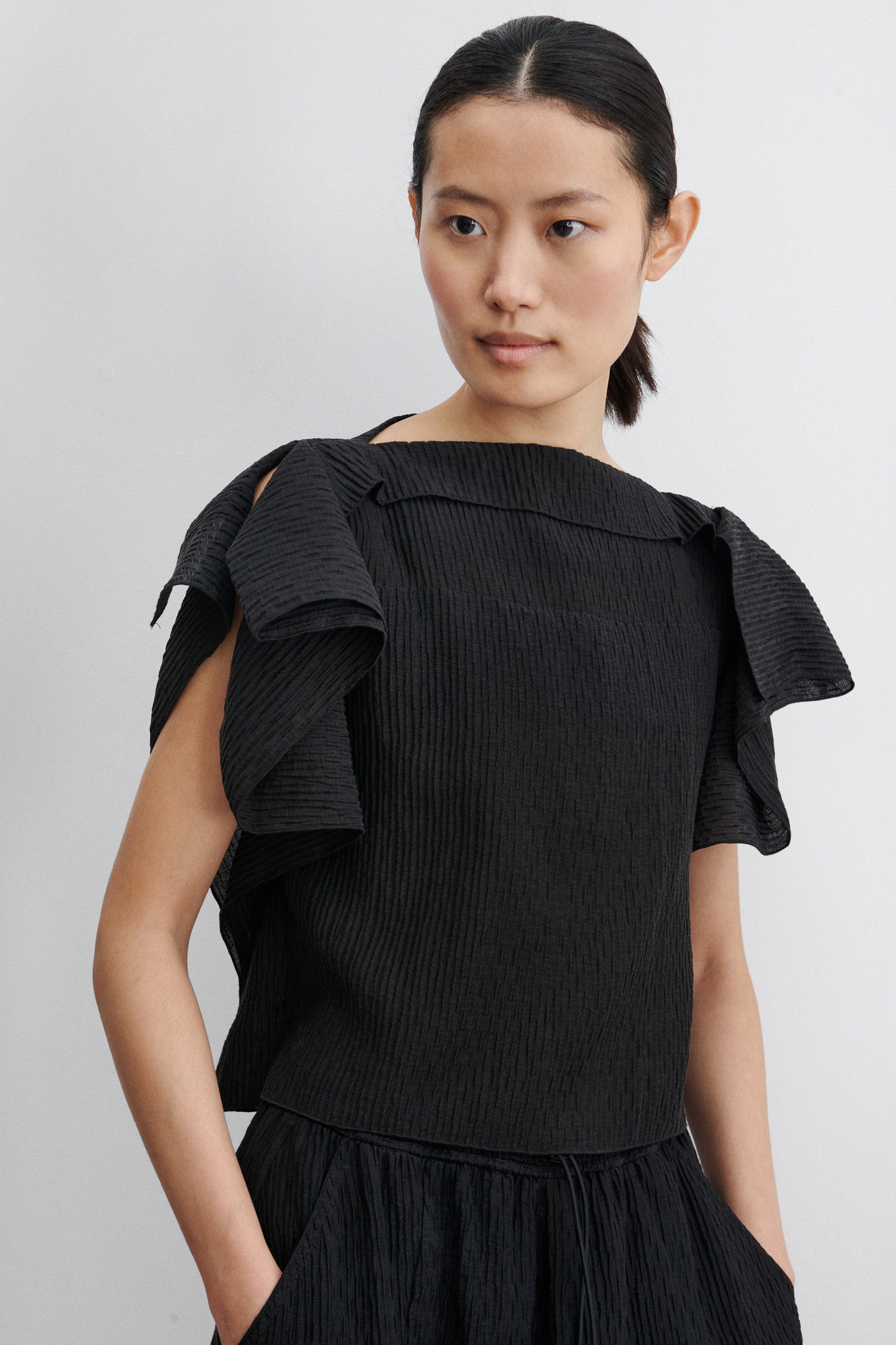 Close-up front view of a woman standing wearing contemporary spring and summer womenswear fashion in a coordinating set by Rachel Comey. The airy Black Rivera Top features softly crinkled fabric, cascading flutter sleeves, and a relaxed, boxy silhouette. Perfectly paired with coordinating Ashbolt Pant for a cohesive look at an Alas Eius Store. Made in the USA. 