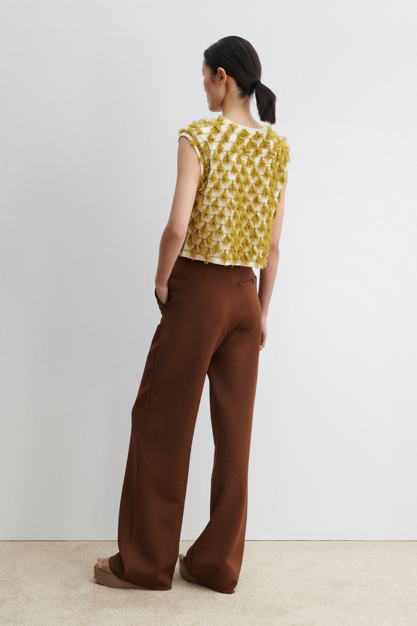 Full length back view of a woman posing wearing retro and vintage inspired fashion in a Rachel Comey Cotton and Baby Alpaca Cream Mustard Hazlitt sleeveless knit top detailed with rows of playful, three-dimensional fringes. Designed with a relaxed, boxy cut and finished with a soft crew neckline at Alas Eius. Made in Peru. 