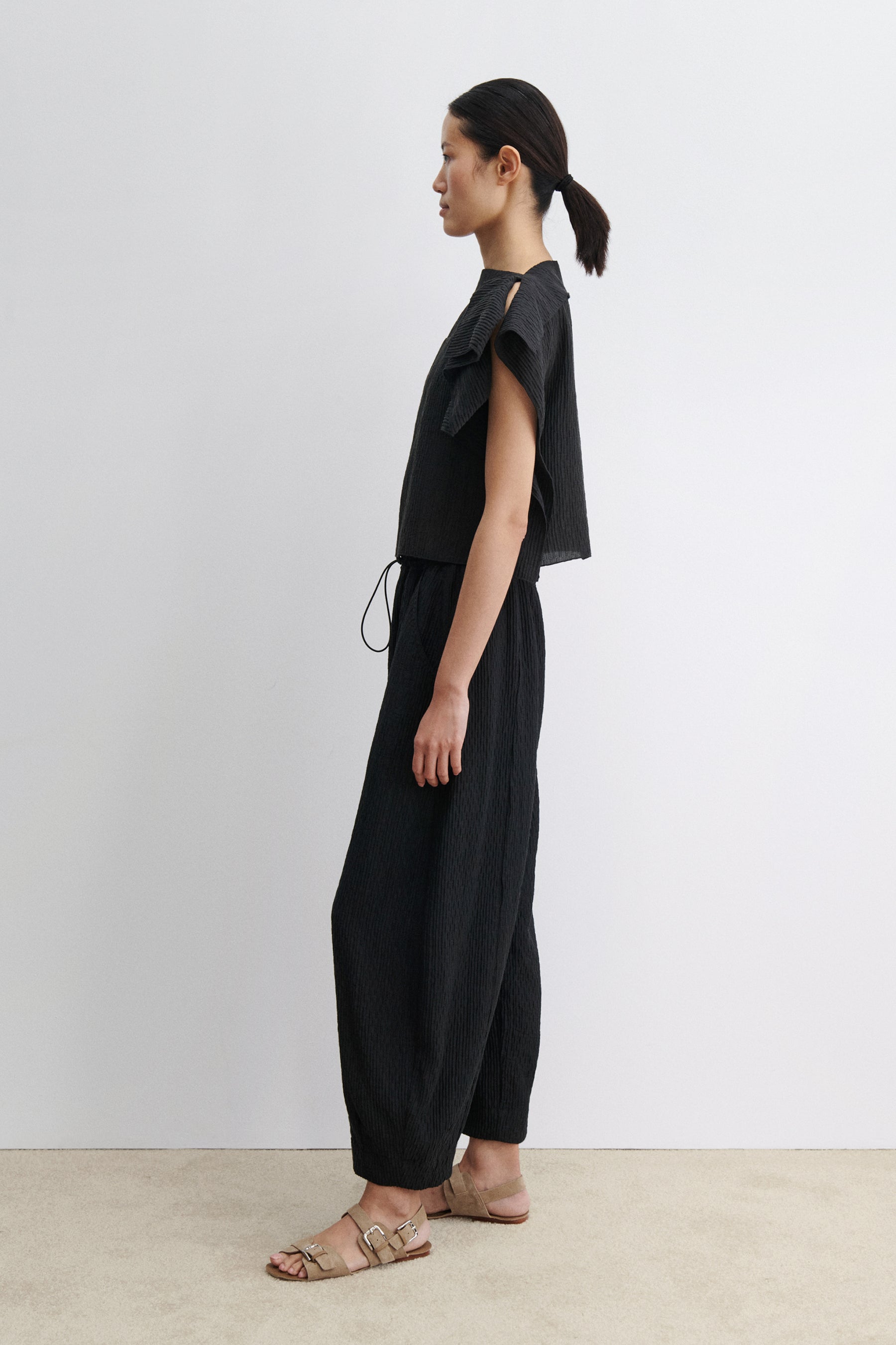 Full length side view of a woman standing wearing contemporary spring and summer womenswear fashion in a coordinating set by Rachel Comey. The airy Black Rivera Top features softly crinkled fabric, cascading flutter sleeves, and a relaxed, boxy silhouette. Perfectly paired with coordinating Ashbolt Pant for a cohesive look at an Alas Eius Store. Made in the USA. 