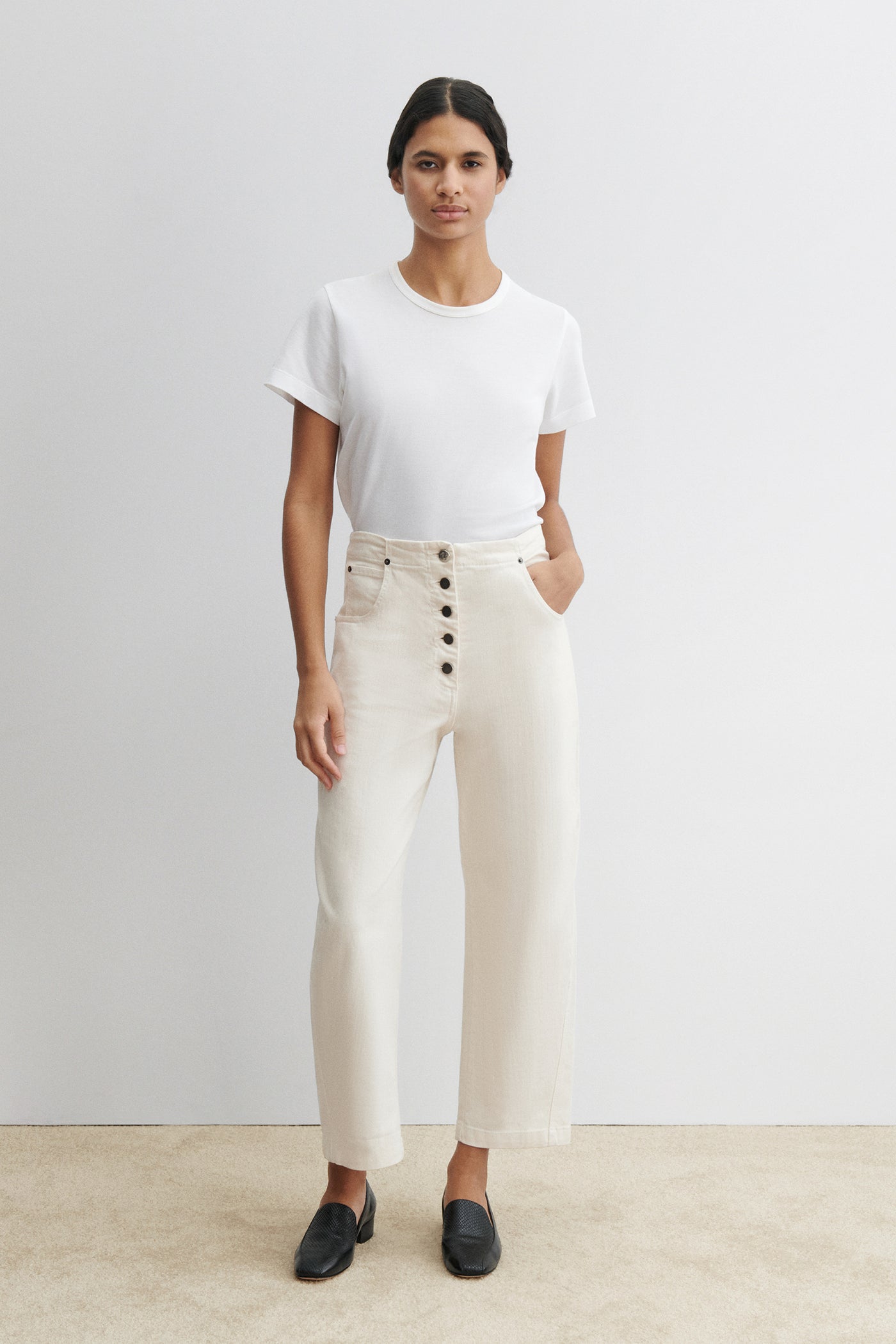 Full length front view of a woman standing wearing chic and elevated spring fashion in a white t-shirt with Rachel Comey White Cotton Elkin Cropped Ankle Pant featuring a 5 button front closure, an adjustable back buckle tab so you can wear at your preferred waistline. Angled darts give the leg a subtle curve at an Alas Eius store. 