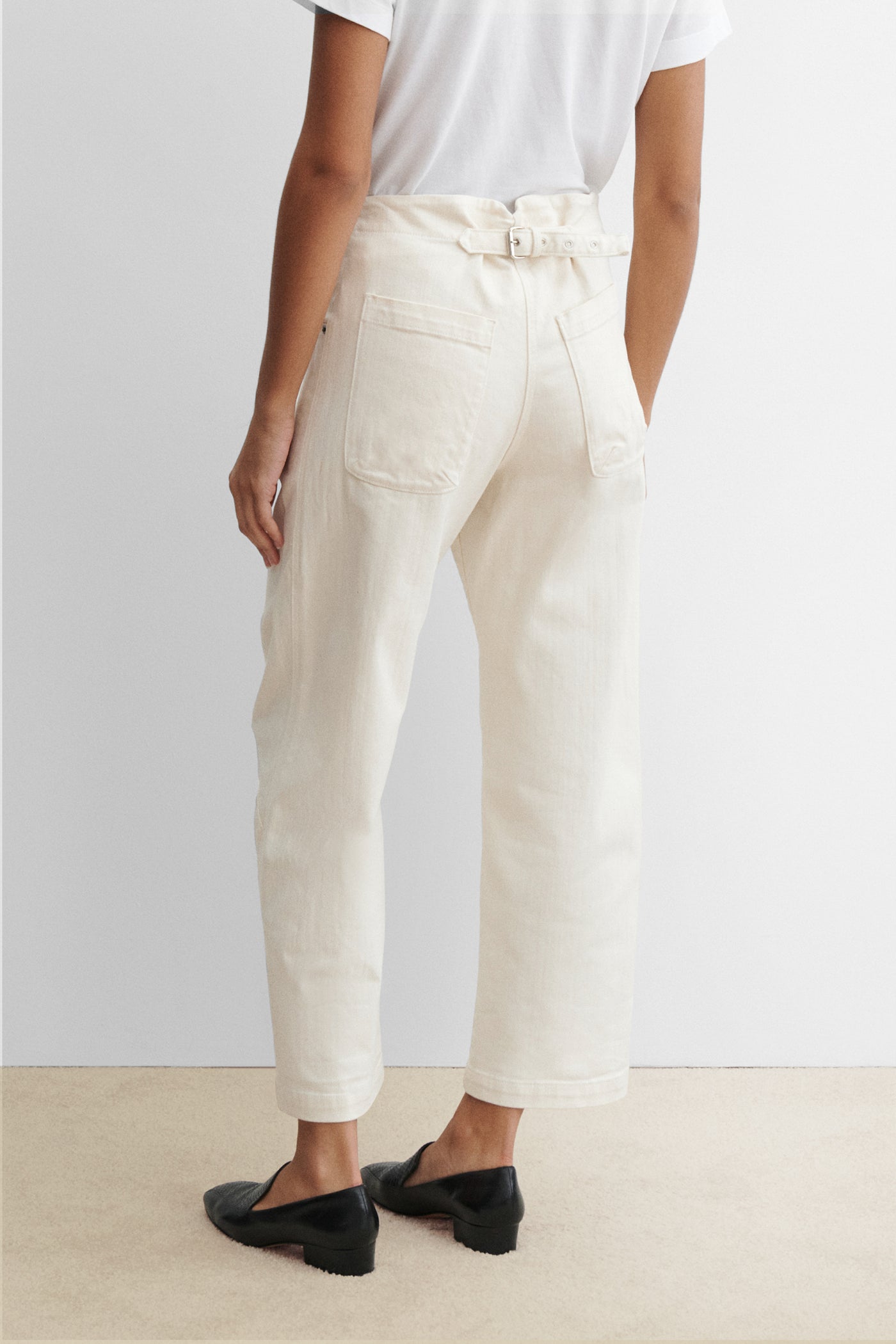 Back view of a woman standing wearing chic and elevated spring fashion in a white t-shirt with Rachel Comey White Cotton Elkin Cropped Ankle Cotton Denim Jeans featuring a 5 button front closure, an adjustable back buckle tab so you can wear at your preferred waistline. Angled darts give the leg a subtle curve at an Alas Eius store. 