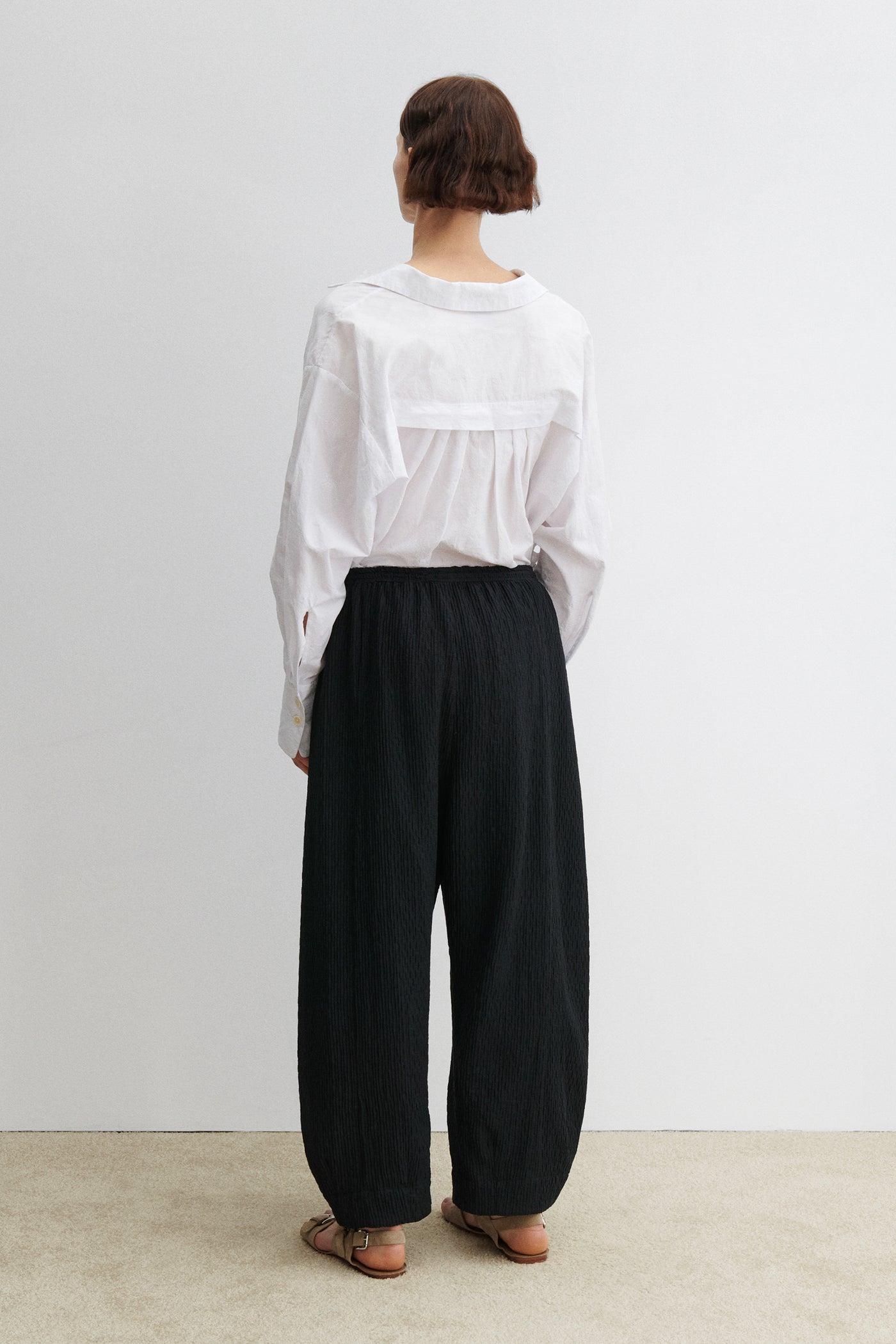 Full length back view of a woman wearing modern minimalist spring summer fashion in the Rachel Comey Black Ashbolt Pant featuring a relaxed wide-leg, elasticated drawstring waistband, two side seam pockets at an Alas Eius store. 