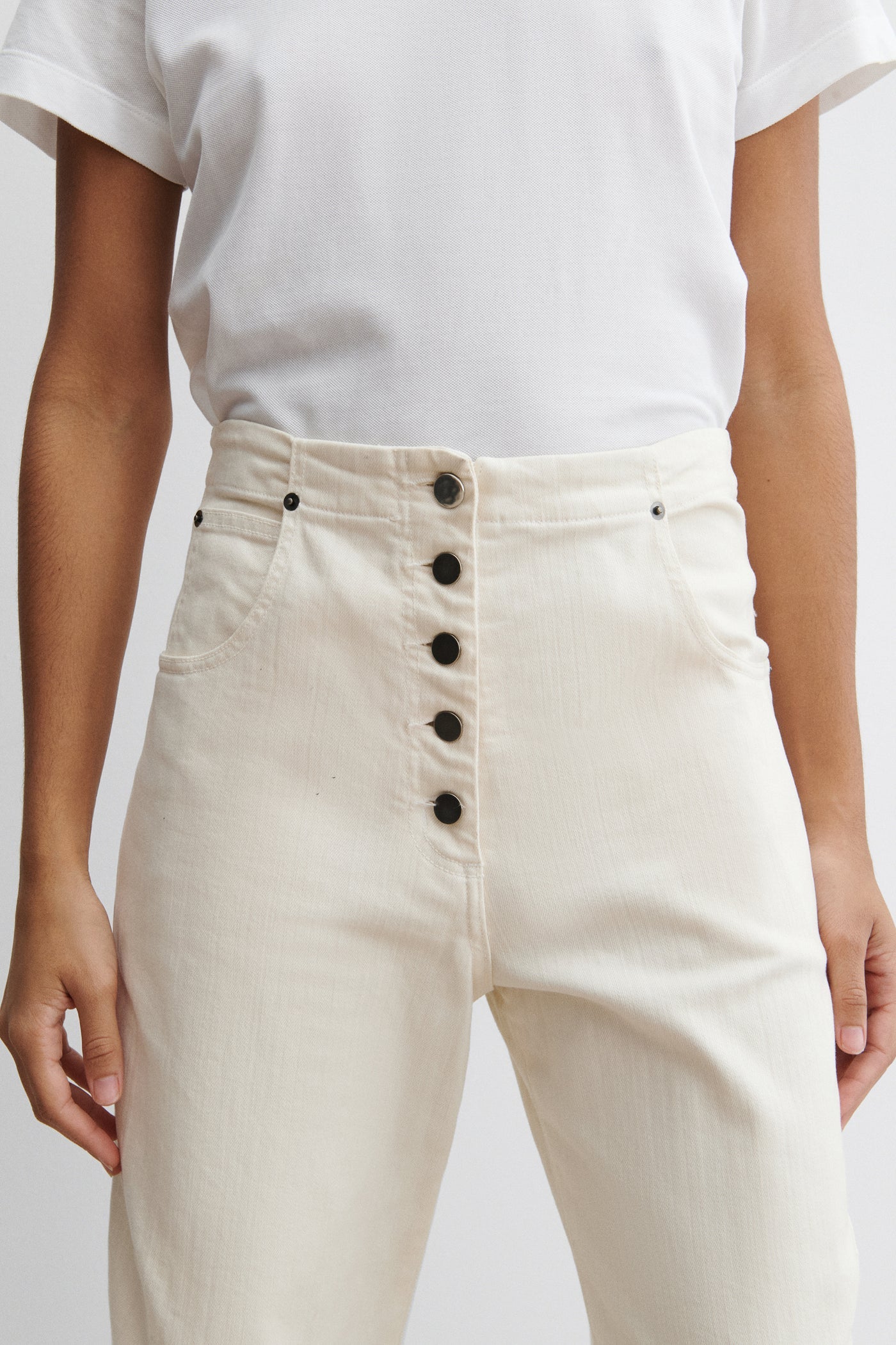 Close-up front view of a woman standing wearing chic and elevated spring fashion in a white t-shirt with Rachel Comey White Cotton Elkin Cropped Ankle Cotton Denim Jeans featuring a 5 button front closure, an adjustable back buckle tab so you can wear at your preferred waistline. Angled darts give the leg a subtle curve at an Alas Eius store. 