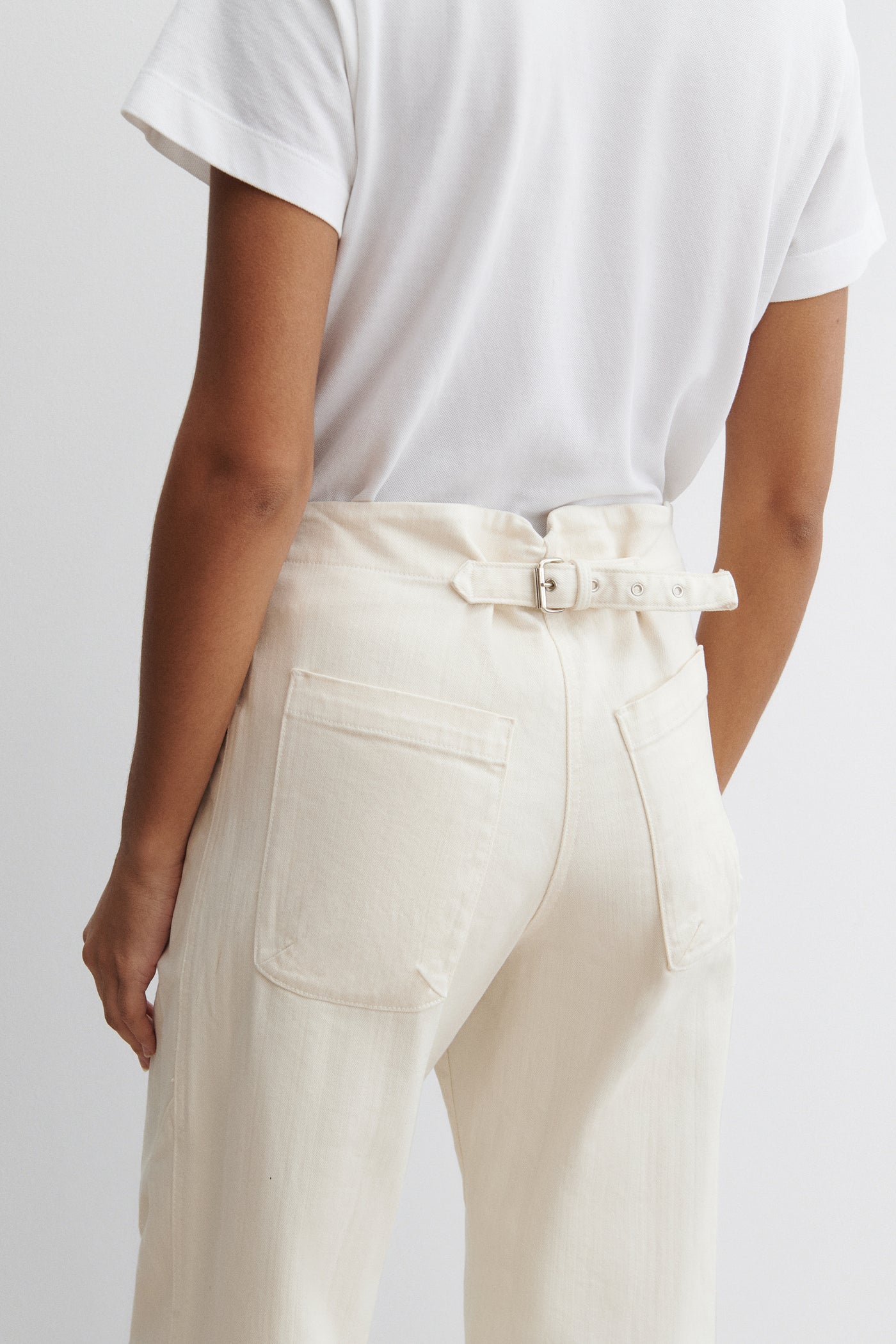 Close-up back view of a woman standing wearing chic and elevated spring fashion in a white t-shirt with Rachel Comey White Cotton Elkin Cropped Ankle Cotton Denim Jeans featuring a 5 button front closure, an adjustable back buckle tab so you can wear at your preferred waistline. Angled darts give the leg a subtle curve at an Alas Eius store. 