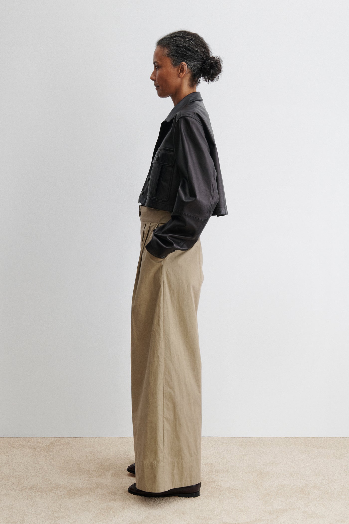 Full length side view of a woman standing wearing chic and versatile spring summer fashion in the Rachel Comey Khaki Coxsone Pant with front pleats, high-waist, and wide leg, cut from signature Organic Crisp cotton. The trouser features side slip pockets, an elastic waistband at the back, a concealed side zipper paired with a cropped faux leather button front jacket at an Alas Eius store. Made in the USA. 