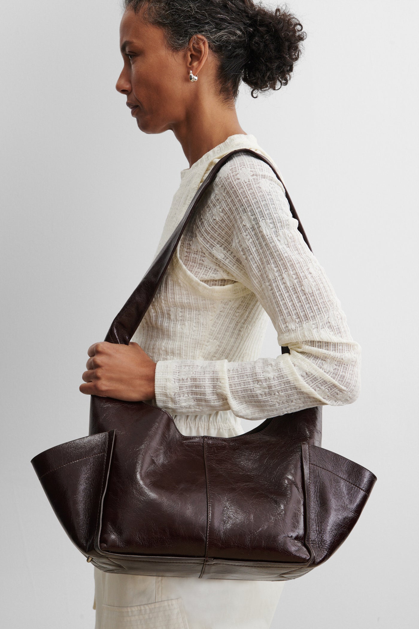 Woman wearing a modern and chic Rachel Comey Chestnut Leather Tool Bag featuring a fixed shoulder strap, extended exterior pockets, and magnetic closure. Can be worn over the shoulder or across the body at an Alas Eius store. Made in Italy. 