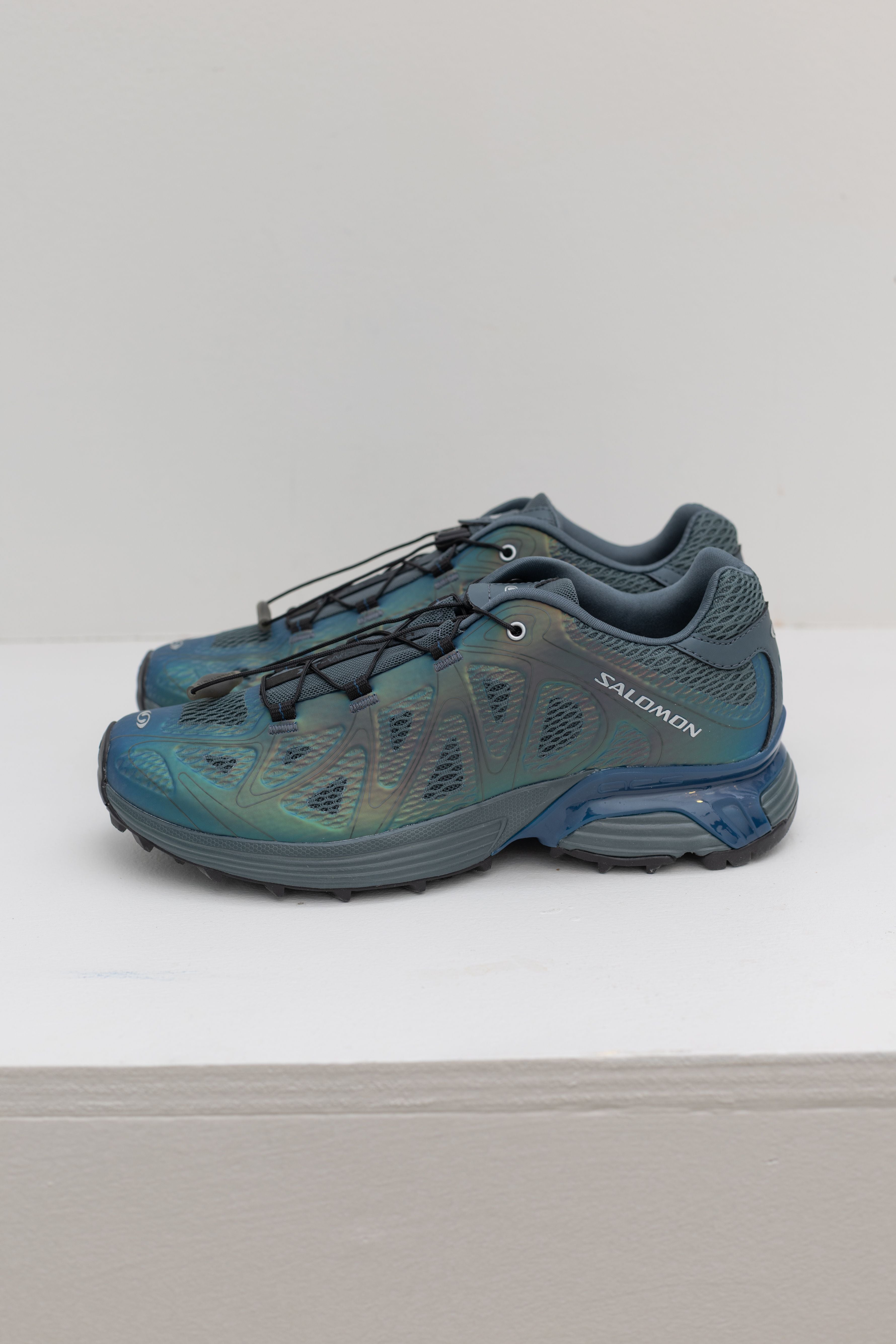Side view of a pair of modern and elevated Salomon XT-Whisper Void Sneakers in a iridescent Dark Slate and Gibraltar Sea colorway featuring quickLACE a minimalistic and strong lace for one-pull tightening and easy-on, easy-off on a white shelf at Alas Eius.