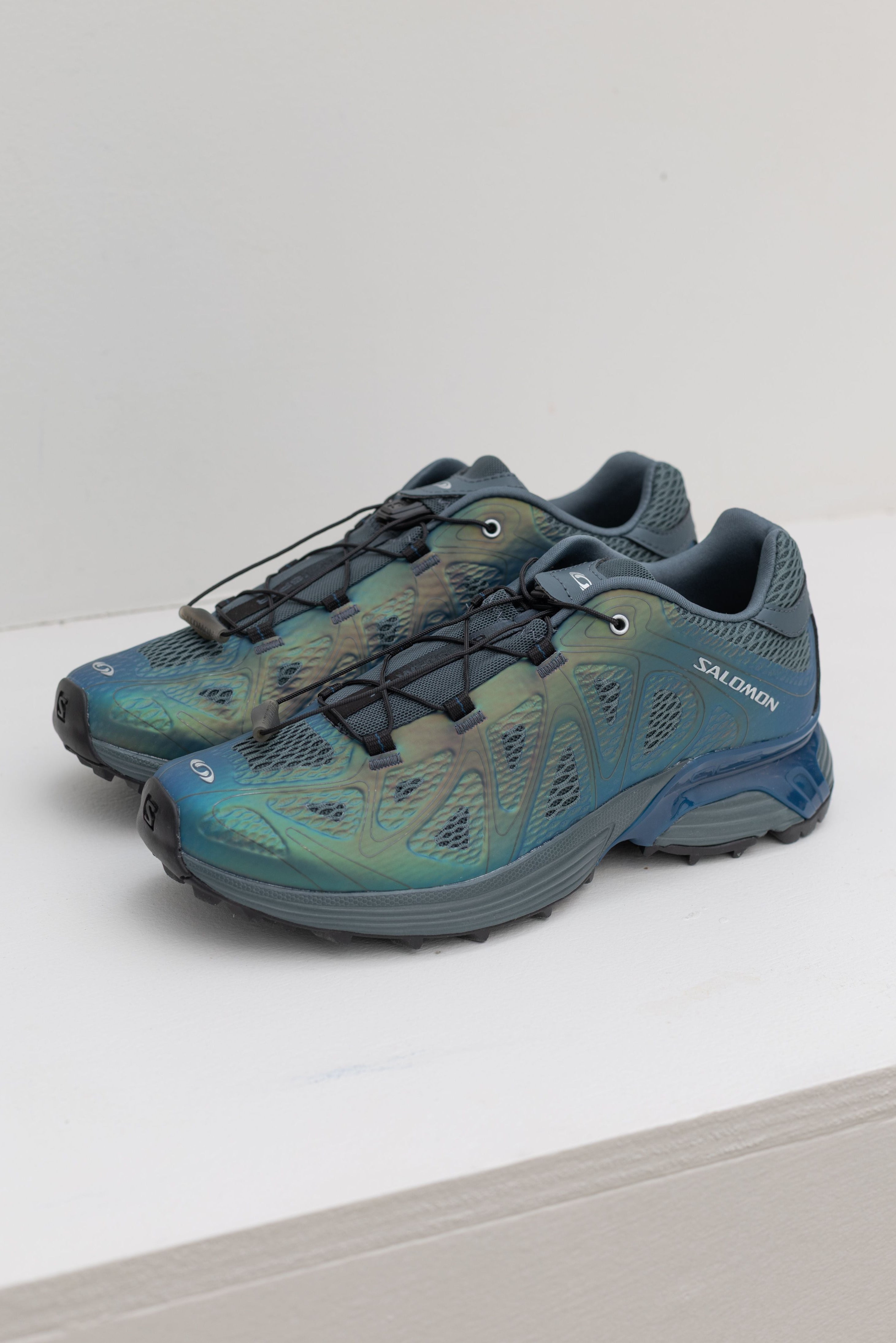 A pair of modern and elevated Salomon XT-Whisper Void Sneakers in a iridescent Dark Slate and Gibraltar Sea colorway featuring quickLACE a minimalistic and strong lace for one-pull tightening and easy-on, easy-off on a white shelf at Alas Eius.