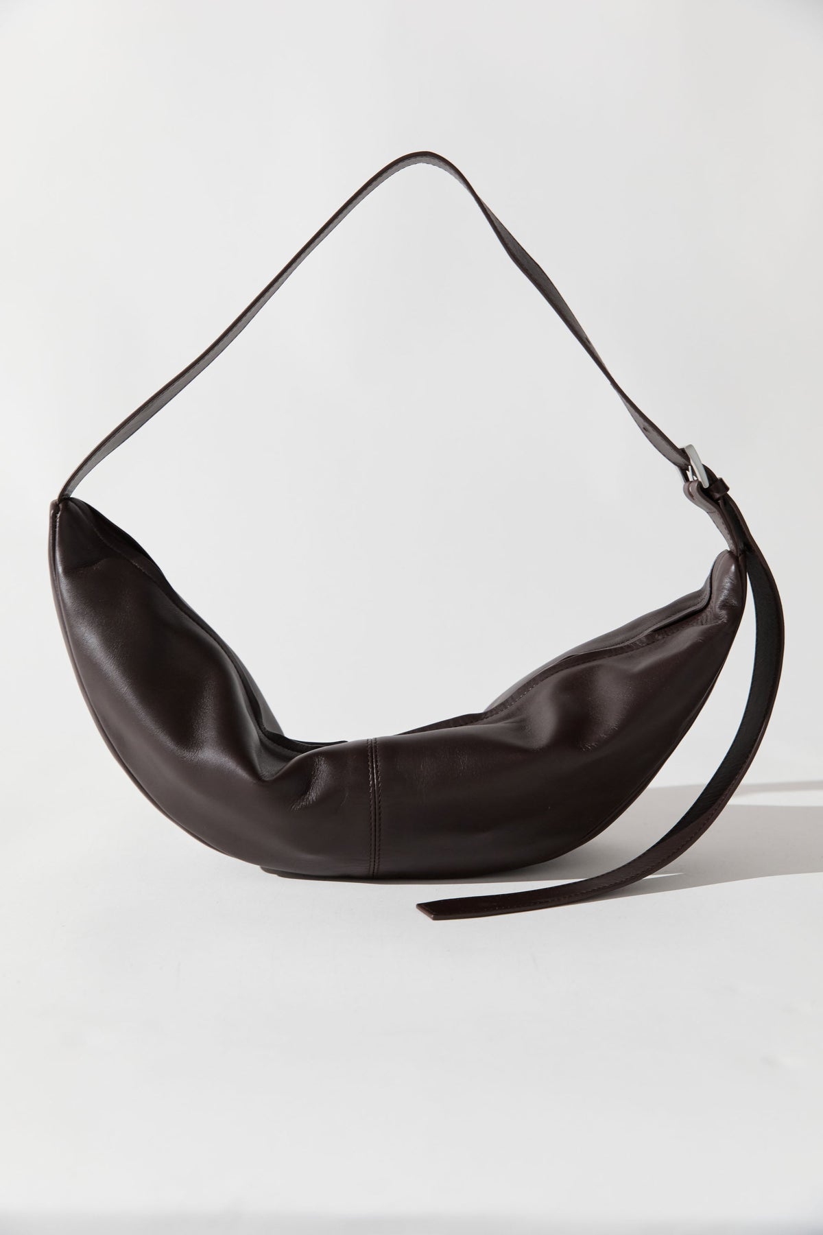 Product view of minimalist design St.Agni Coffee brown leather Adjustable strap Slim Crescent shaped Bag at Alas Eius