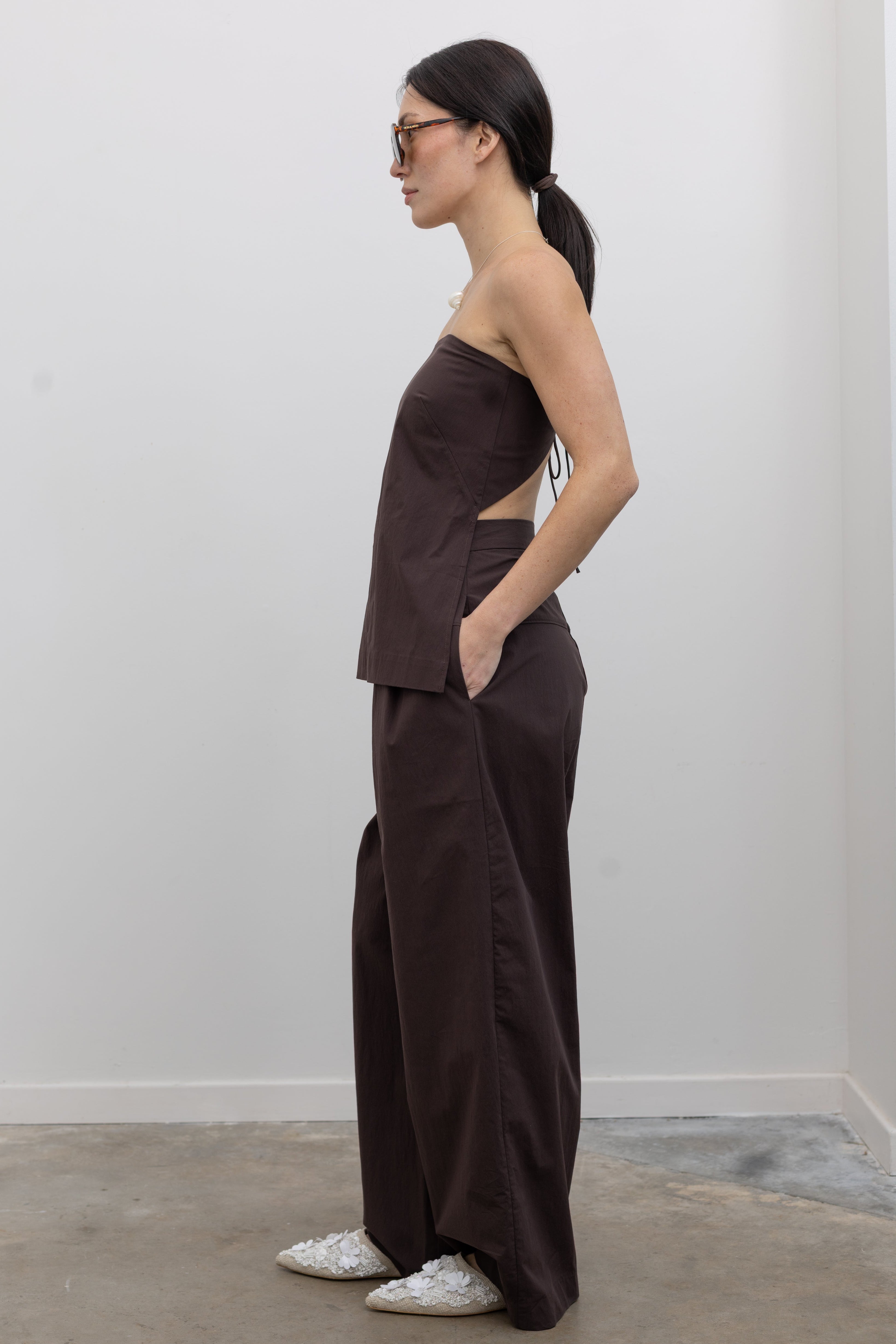 Full side view of a woman standing wearing Spring and Summer fashion in the St. Agni Bark Brown Relaxed Pleat Pants featuring a mid-rise elastic waist and a gently tapered leg paired with its coordinating Bark Brown Strapless Rouleau tie back top at to create the perfect set Alas Eius.