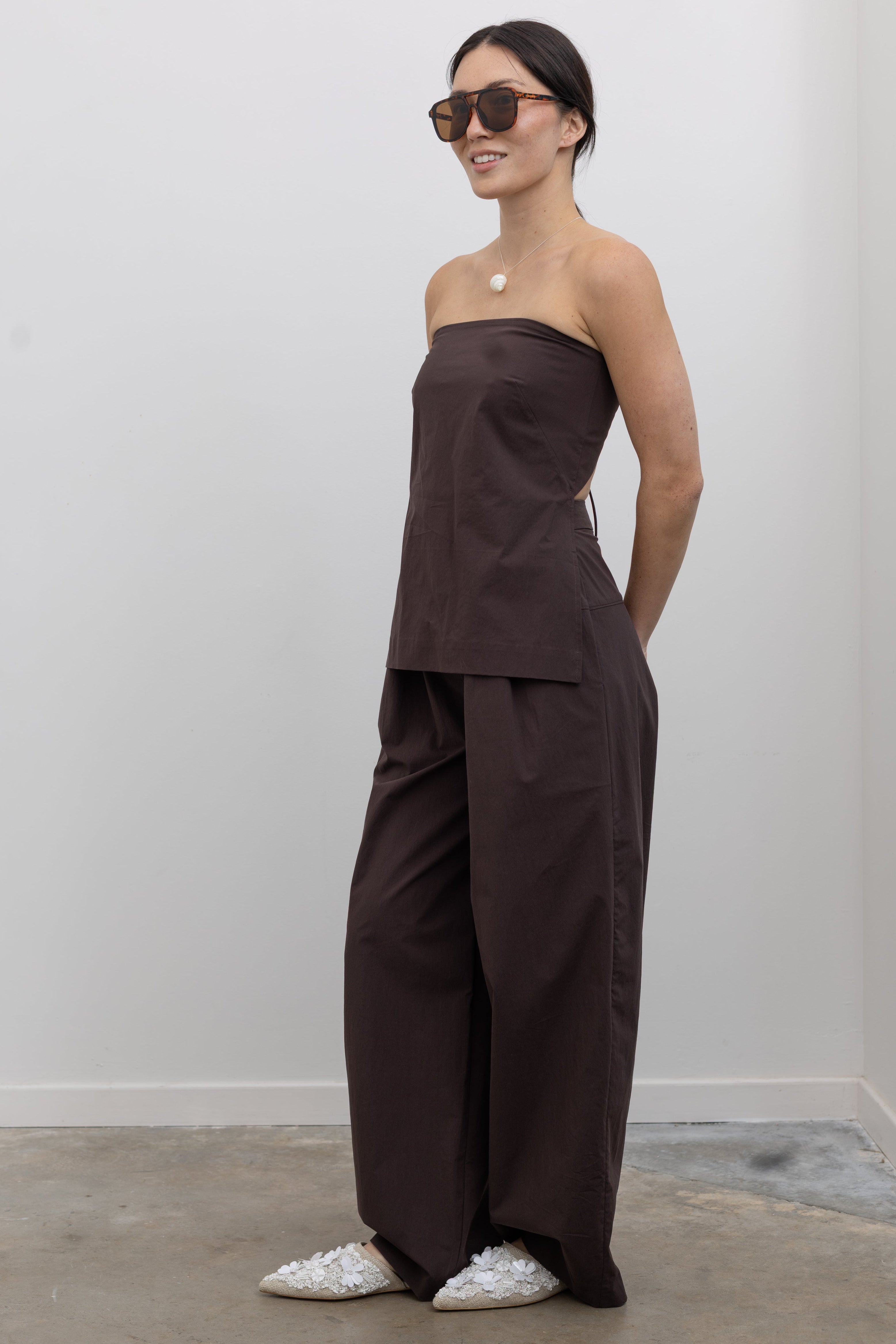 3/4 Full length view of a woman wearing spring fashion in a St. Agni Bark Brown Strapless Rouleau Back Tie Top with its coordinating pants to create the perfect set at Alas Eius.