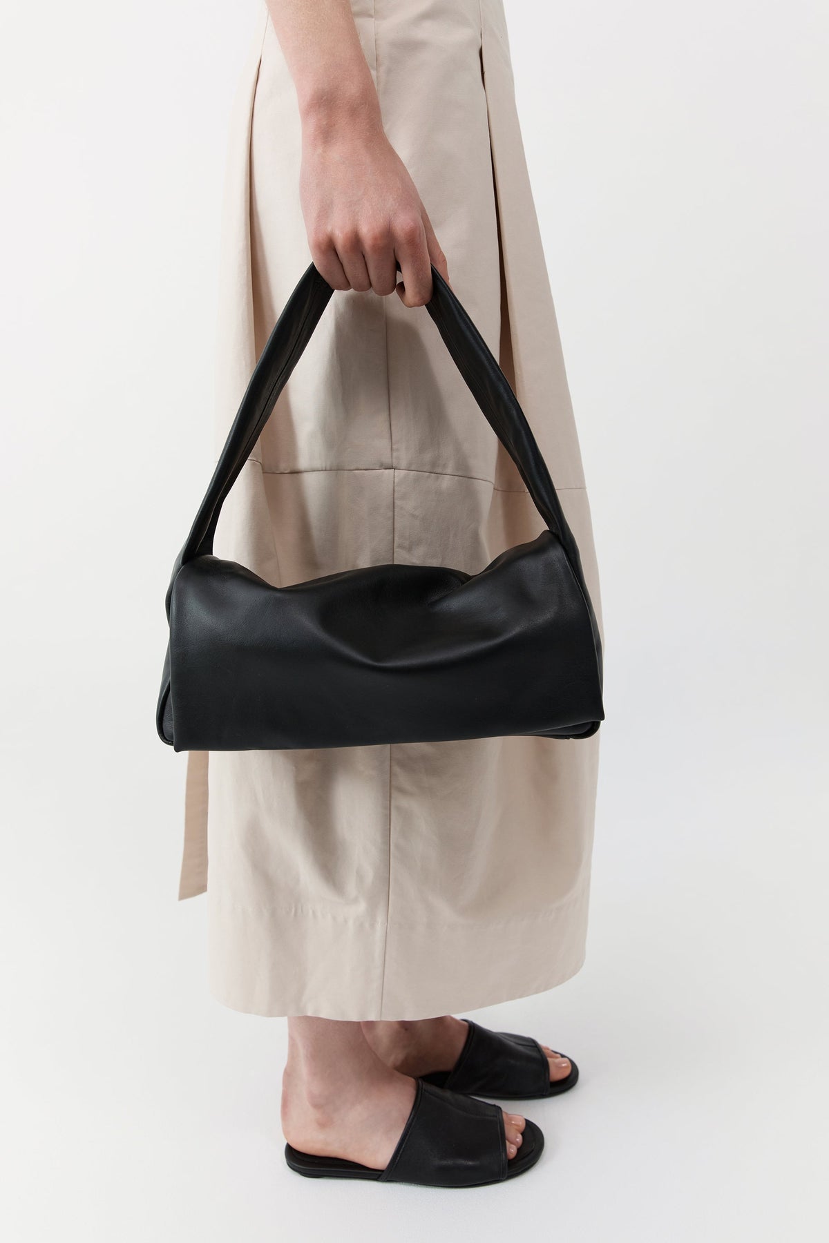 Woman standing holding a St. Agni black leather soft slim satchel bag which is lined with cotton canvas and features a magnetic closure at Alas Eius.