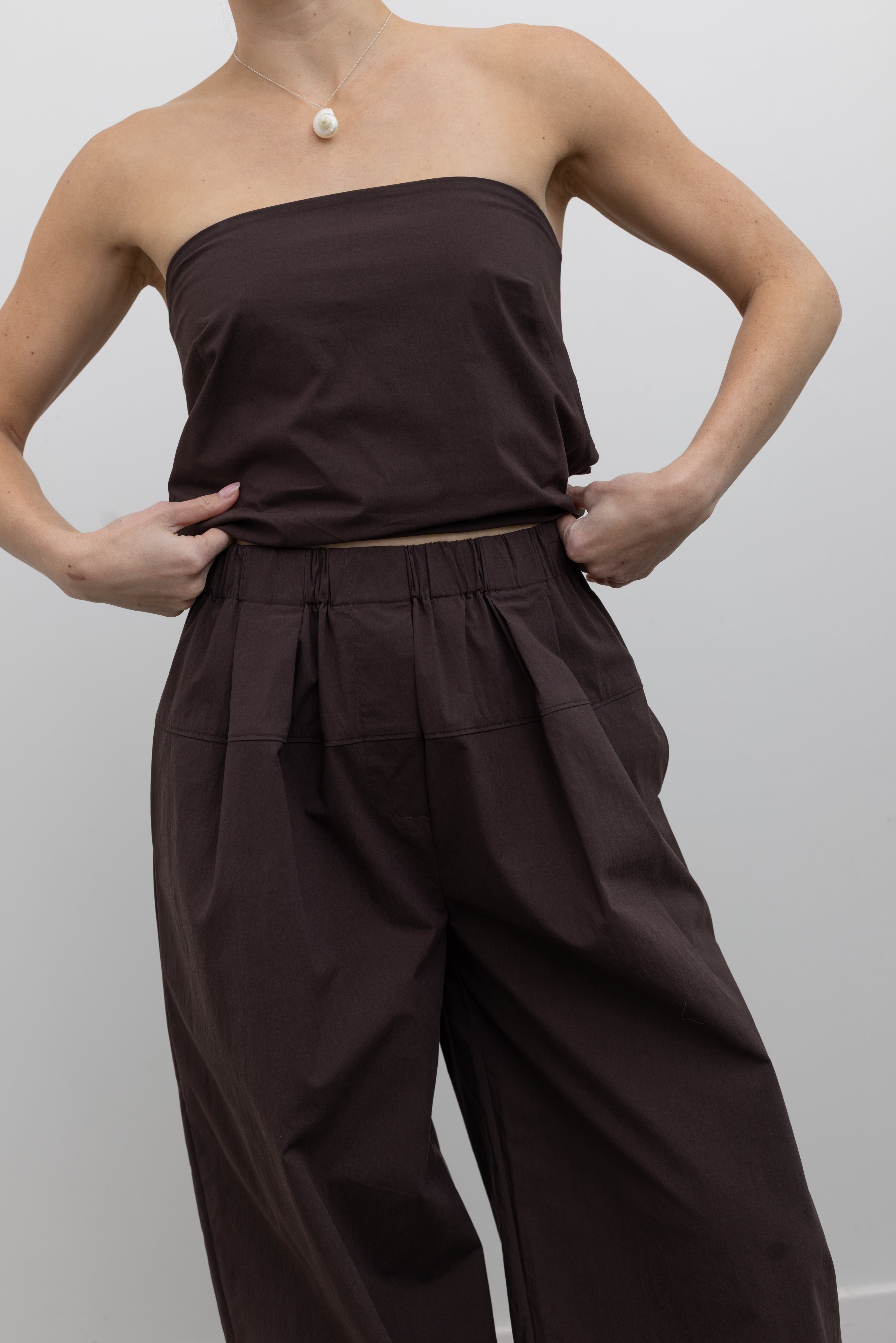 Close-up detailed front view of a woman standing wearing Spring and Summer fashion in the St. Agni Bark Brown Relaxed Pleat Pants featuring a mid-rise elastic waist and a gently tapered leg paired with its coordinating strapless tie back top at Alas Eius.
