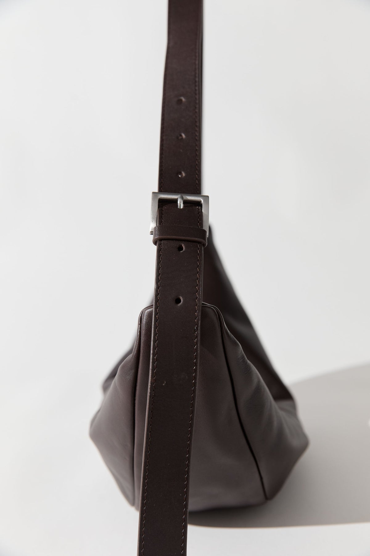 Close-up side view of a St.Agni label Coffee brown leather Adjustable strap Slim Crescent shaped Bag at Alas Eius