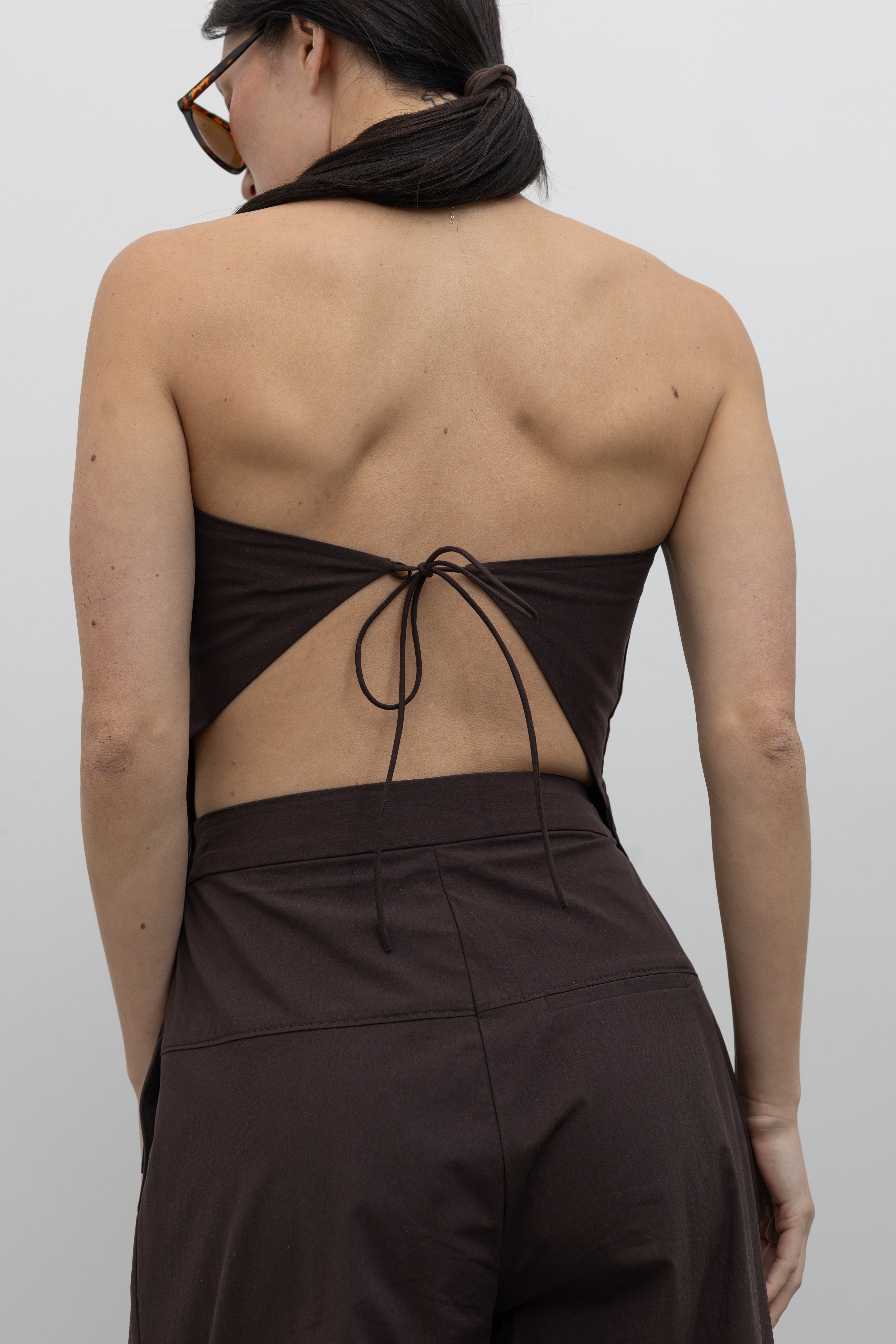 Close-up detailed back view of a woman standing wearing Spring and Summer fashion in the St. Agni Bark Brown Relaxed Pleat Pants featuring a mid-rise elastic waist and a gently tapered leg paired with its coordinating Bark Brown Strapless Rouleau tie back top at to create the perfect set Alas Eius.