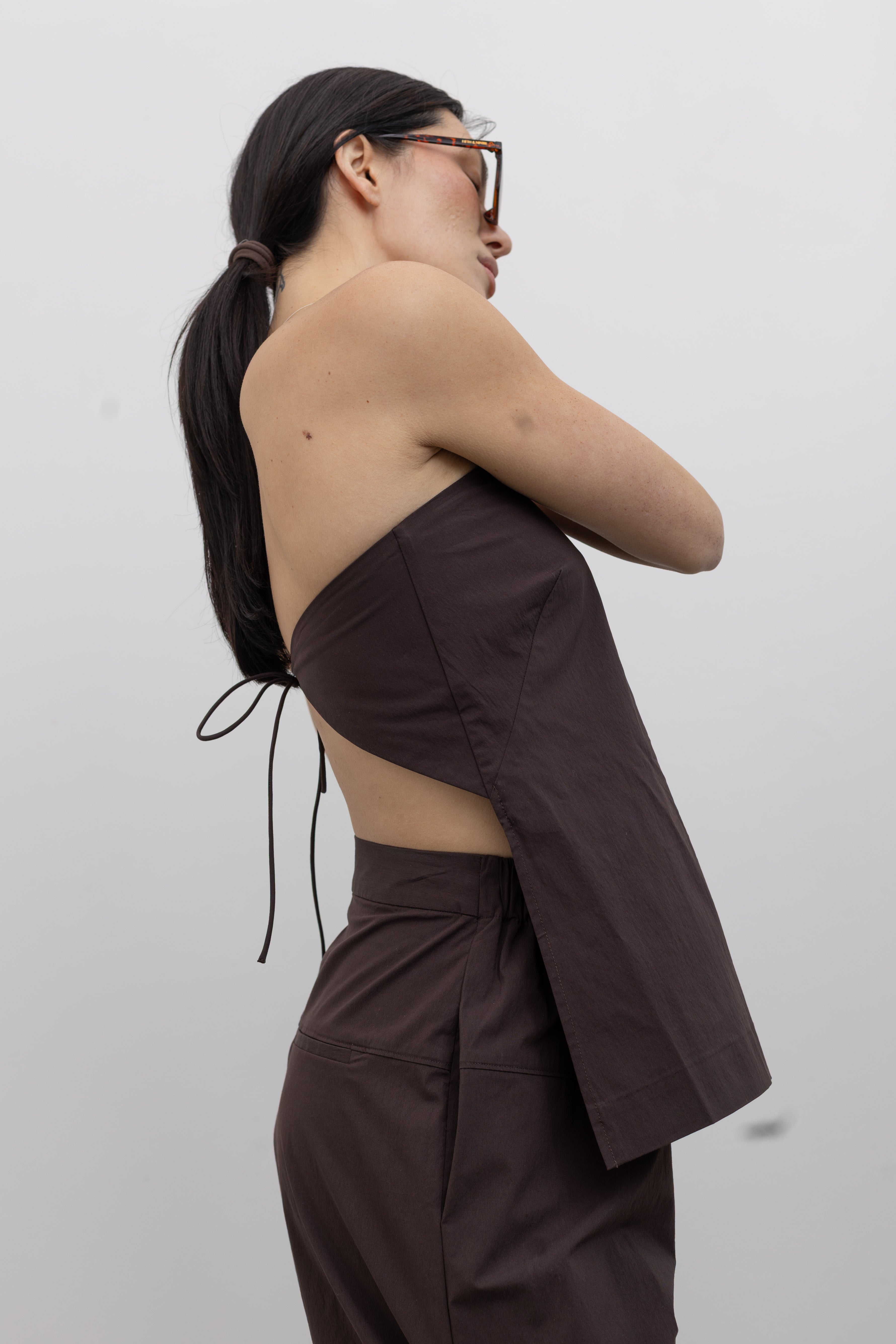 Side view of a woman standing wearing Spring and Summer fashion in the St. Agni Bark Brown Relaxed Pleat Pants featuring a mid-rise elastic waist and a gently tapered leg paired with its coordinating Bark Brown Strapless Rouleau tie back top at to create the perfect set Alas Eius.