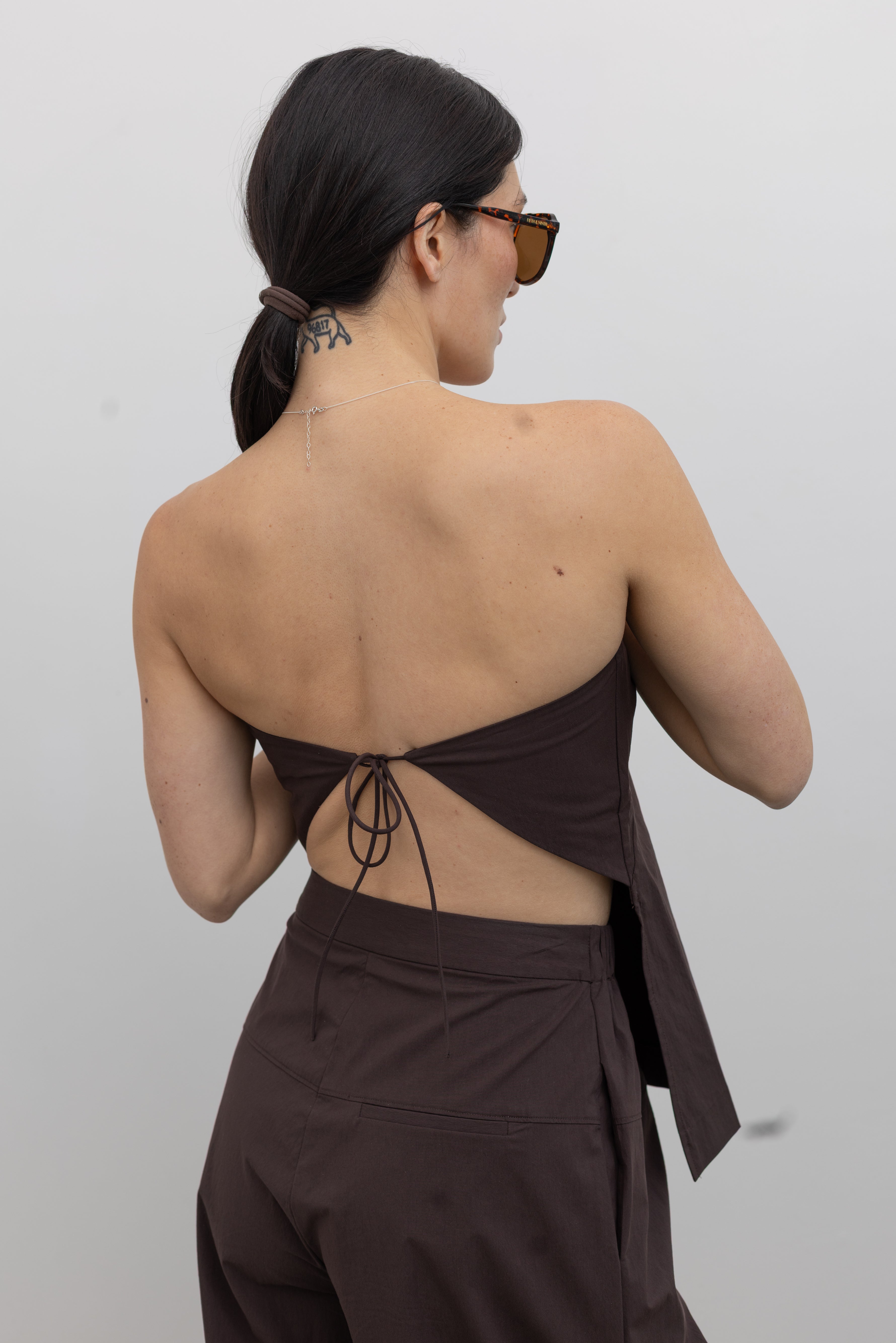 Back view of a woman standing wearing Spring and Summer fashion in the St. Agni Bark Brown Relaxed Pleat Pants featuring a mid-rise elastic waist and a gently tapered leg paired with its coordinating Bark Brown Strapless Rouleau tie back top at to create the perfect set Alas Eius.
