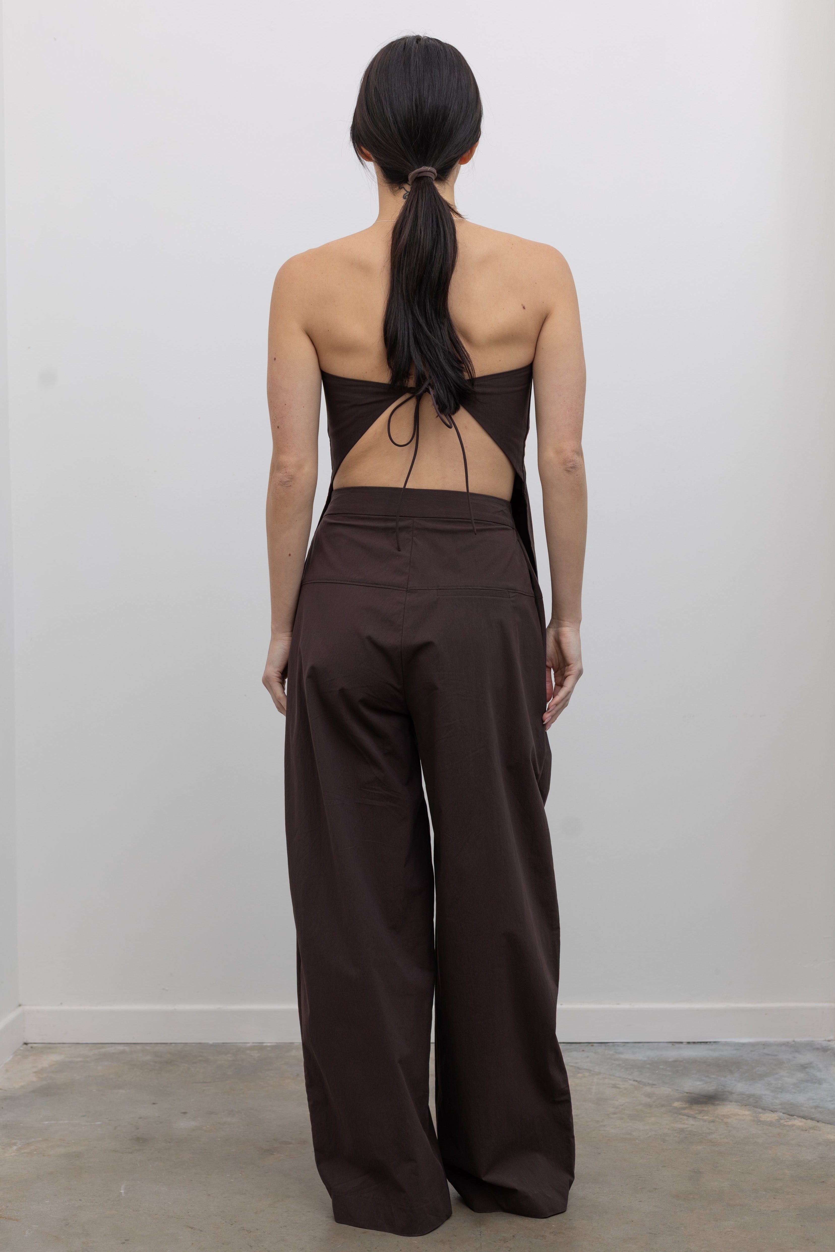 Back view of a woman standing wearing Spring and Summer fashion in the St. Agni Bark Brown Relaxed Pleat Pants featuring a mid-rise elastic waist and a gently tapered leg paired with its coordinating Bark Brown Strapless Rouleau tie back top at to create the perfect set Alas Eius.