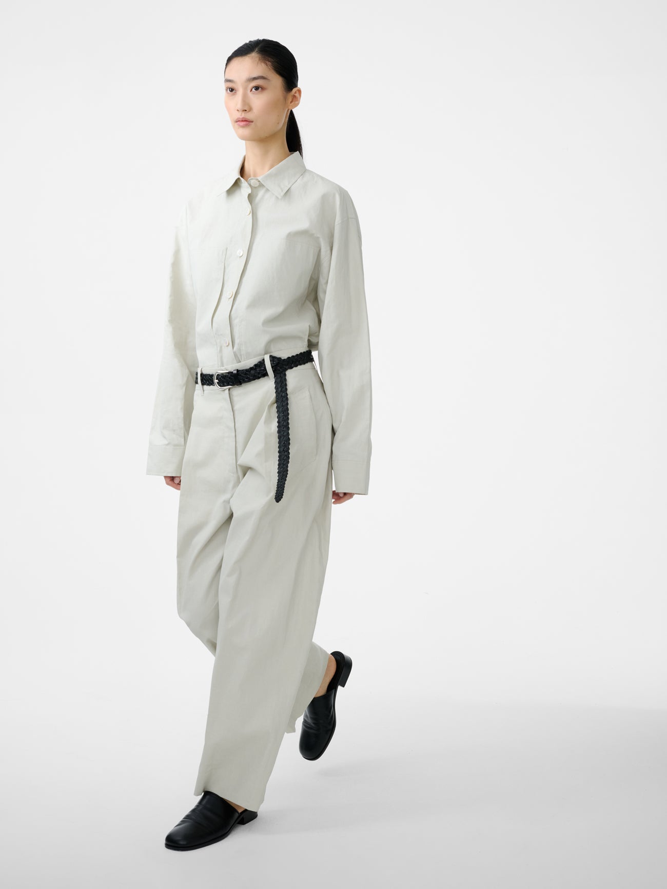 Woman standing wearing modern minimalist spring fashion in the Studio Nicholson Bone Minos Pant cut from coated linen, fitted with broad welt pockets and single reverse pleats, accentuating the volume in the leg at an Alas Eius store.