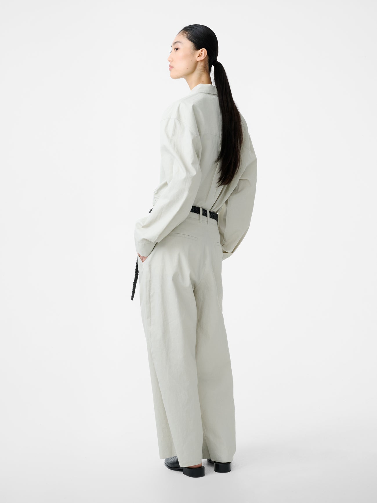 Full length back view of a woman standing wearing modern minimalist spring fashion in the Studio Nicholson Bone Minos Pant cut from coated linen, fitted with broad welt pockets and single reverse pleats, accentuating the volume in the leg at an Alas Eius store.