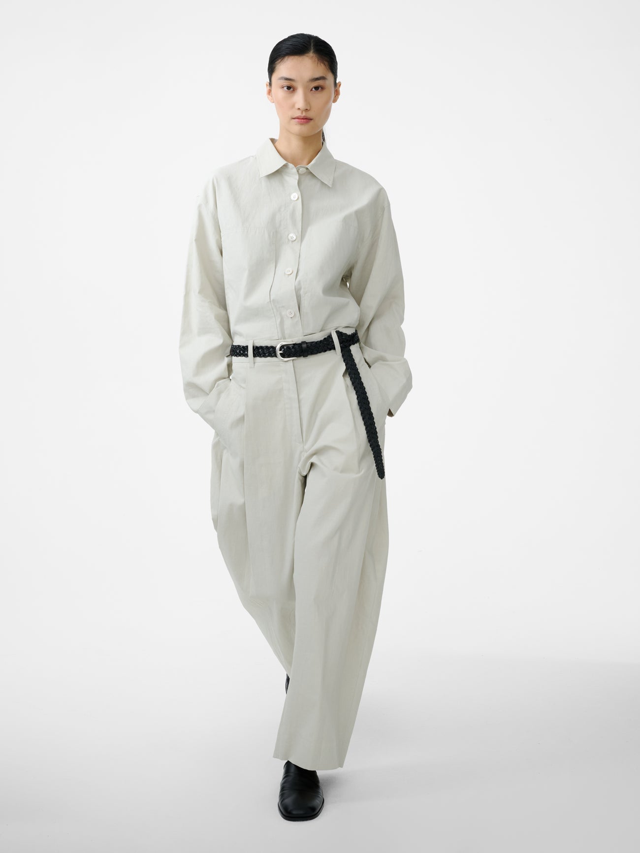 Full length front view of a woman in motion wearing an elevated classic spring fashion staple in the Studio Nicholson Bone Minos Pant cut from coated linen, fitted with broad welt pockets and single reverse pleats, accentuating the volume in the leg at an Alas Eius store.