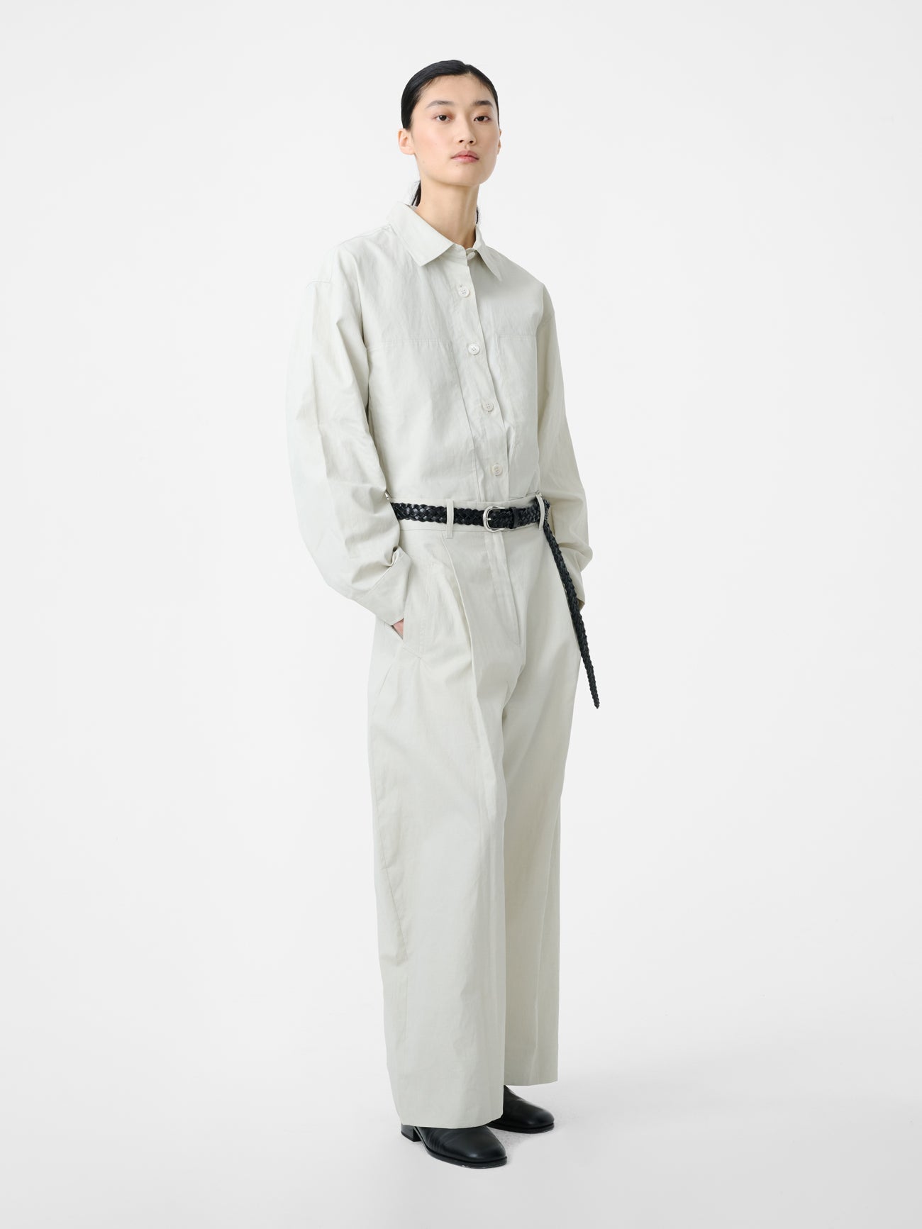 Woman standing wearing modern minimalist spring fashion in the Studio Nicholson Bone Minos Pant Minos cut from coated linen, fitted with broad welt pockets and single reverse pleats, accentuating the volume in the leg at an Alas Eius store.