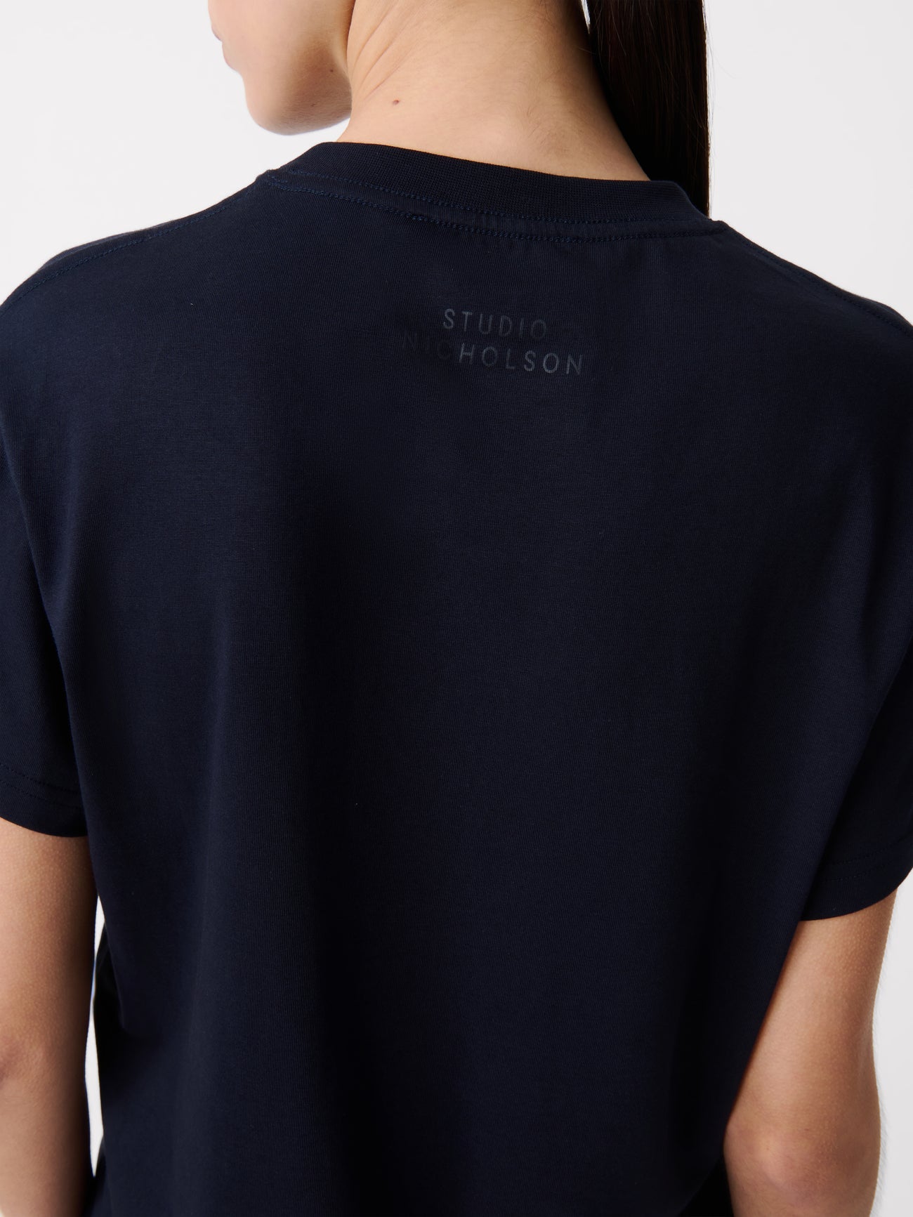 Close-up back view of a woman standing wearing an elevated essential spring fashion staple in a Studio Nicholson Darkest Navy Marine T-shirt featuring a classic regular fit and crew neck paired with dark wash denim barrel leg jeans at Alas Eius.