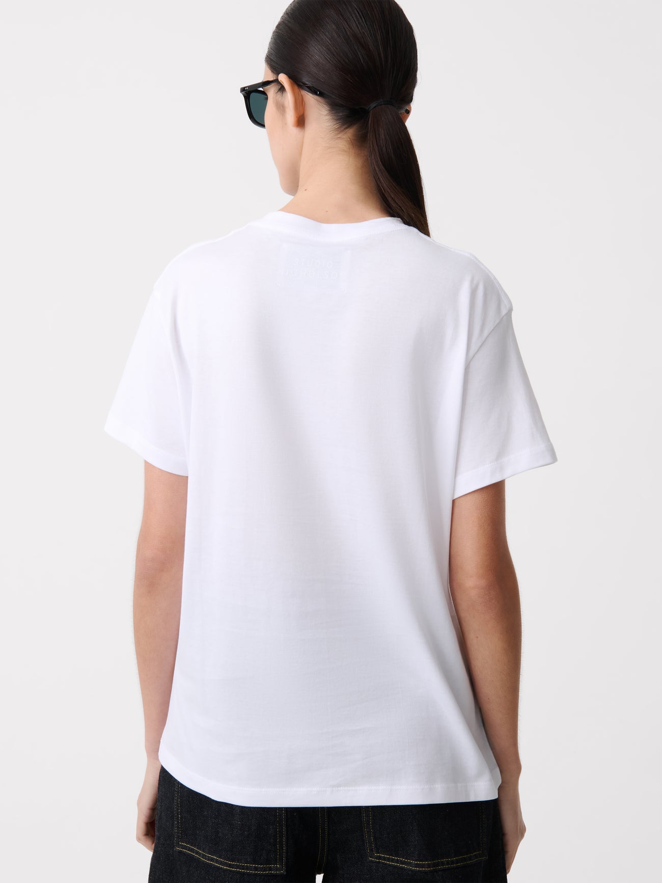 Close-up back view of a woman standing wearing an elevated essential spring fashion staple in a Studio Nicholson Off White Marine T-shirt featuring a classic regular fit and crew neck paired with dark wash denim barrel leg jeans at Alas Eius.