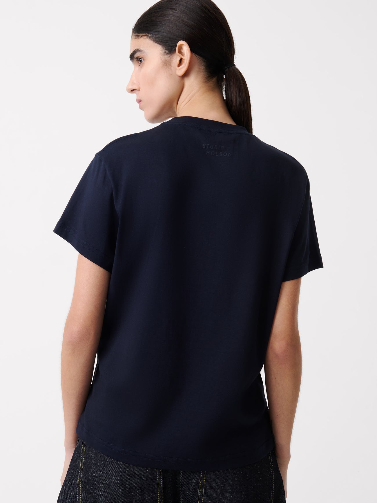 Close-up back view of a woman standing wearing an elevated essential spring fashion staple in a Studio Nicholson Darkest Navy Marine T-shirt featuring a classic regular fit and crew neck paired with dark wash denim barrel leg jeans at Alas Eius.