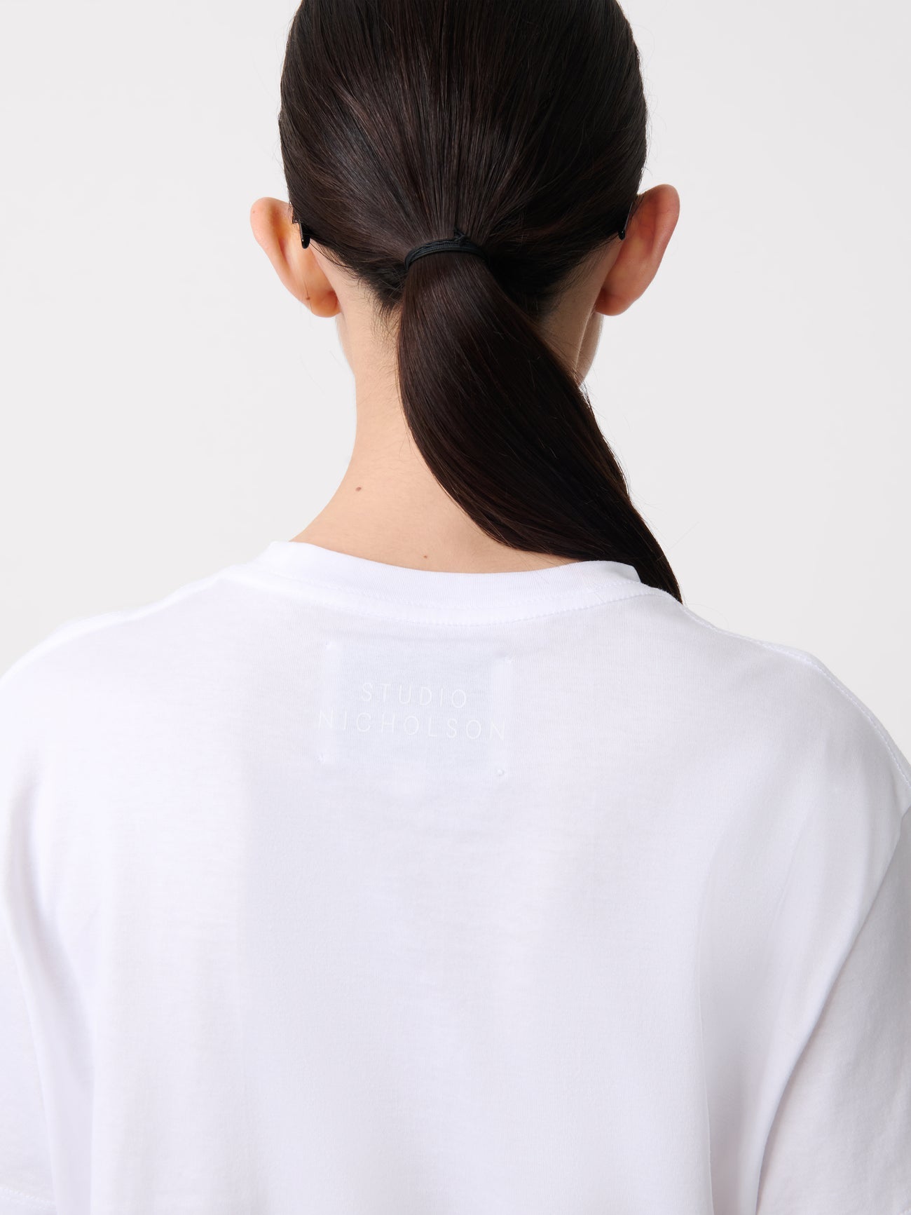 Close-up back view of a woman standing wearing an elevated essential spring fashion staple in a Studio Nicholson Off White Marine T-shirt featuring a classic regular fit and crew neck paired at Alas Eius.