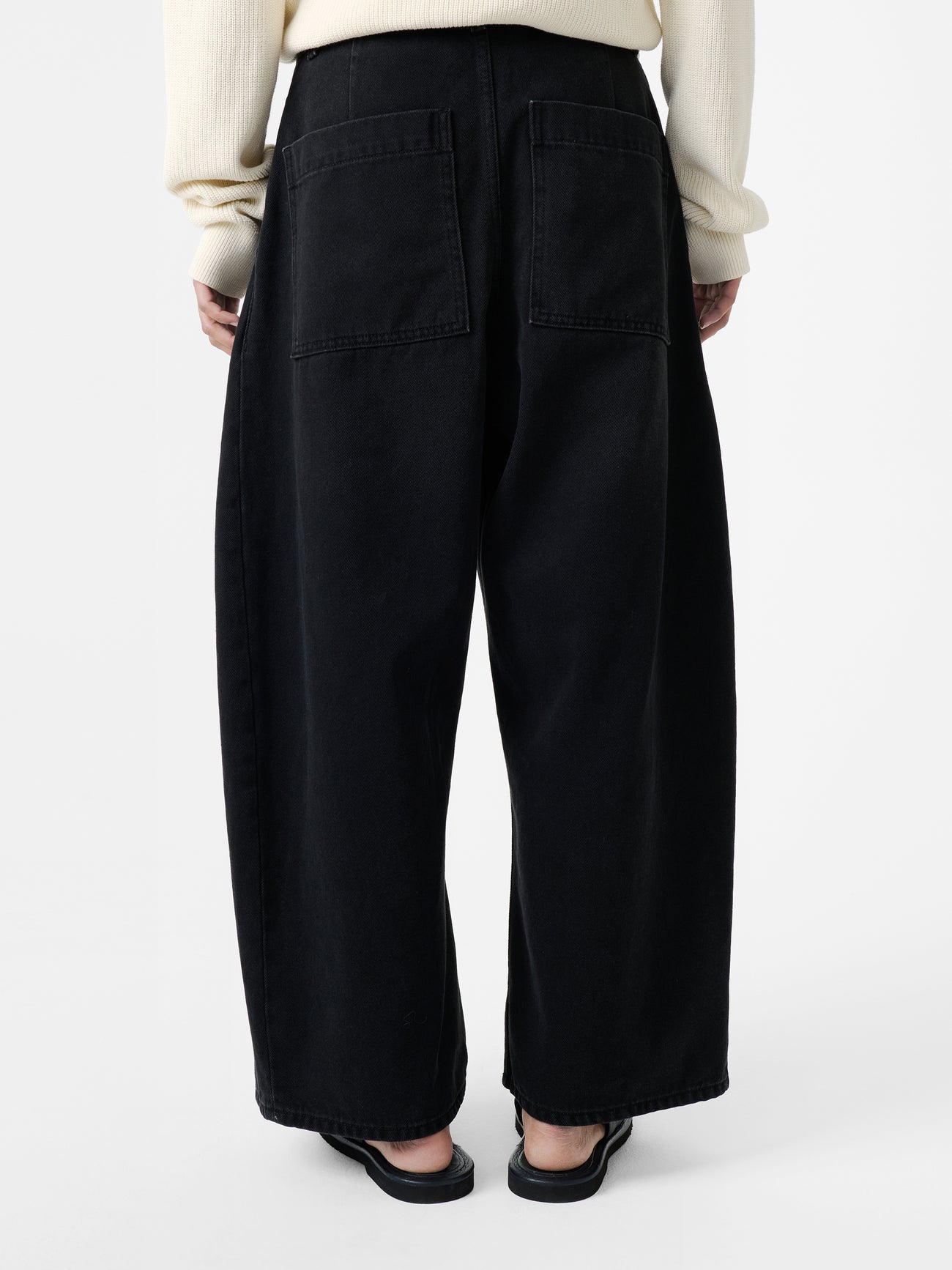 Close-up back view of a woman wearing elevated casual weekend essential style in the Studio Nicholson Washed Black Chalco Wide Crop Denim Pant that are cut wide and cropped, with a curved leg and dropped crotch at Alas Eius. They are constructed from a denim that’s been woven from regenerative cotton and made in the UK. 