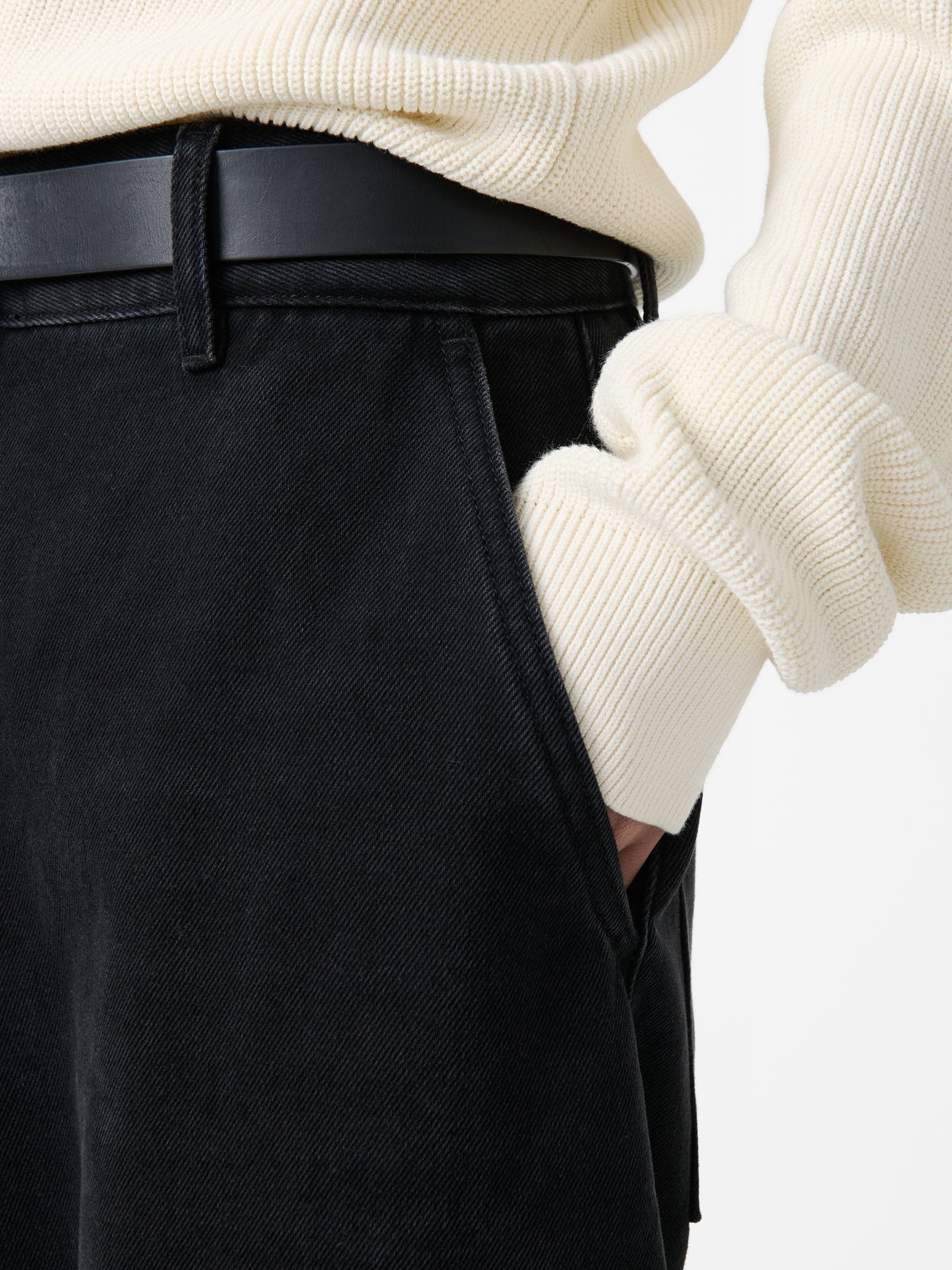 Close-up view of a woman with her hand in the pocket of the Studio Nicholson Washed Black Chalco Wide Crop Denim Pant that are cut wide and cropped, with a curved leg and dropped crotch at Alas Eius. They are constructed from a denim that’s been woven from regenerative cotton and made in the UK. 