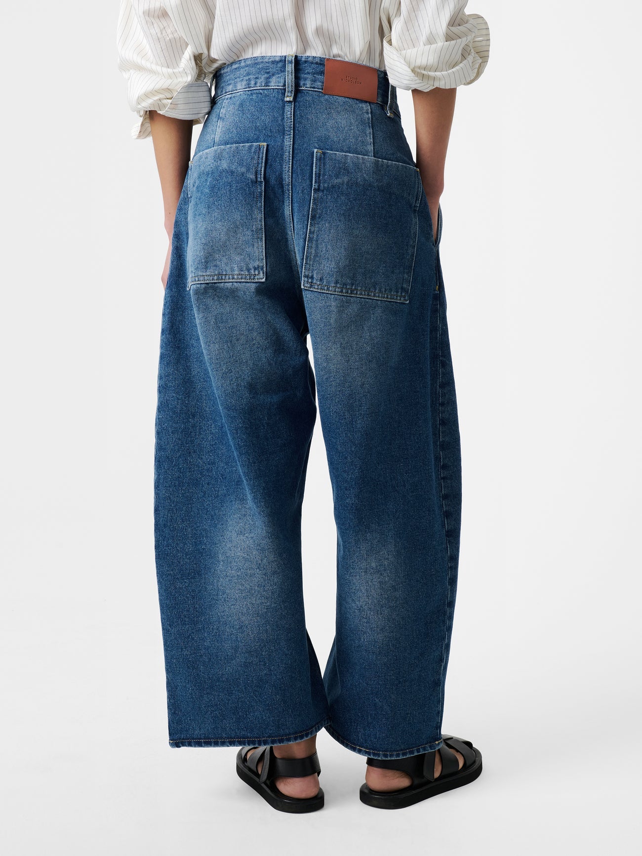 Close-up back view of a woman standing wearing an elevated classic and wardrobe staple in the Studio Nicholson Worn Wash Indigo Chalco Wide Crop Denim Jeans that are cut wide and cropped, with a curved leg and dropped crotch at Alas Eius. They are constructed from a denim that’s been woven from regenerative cotton and made in the UK.
