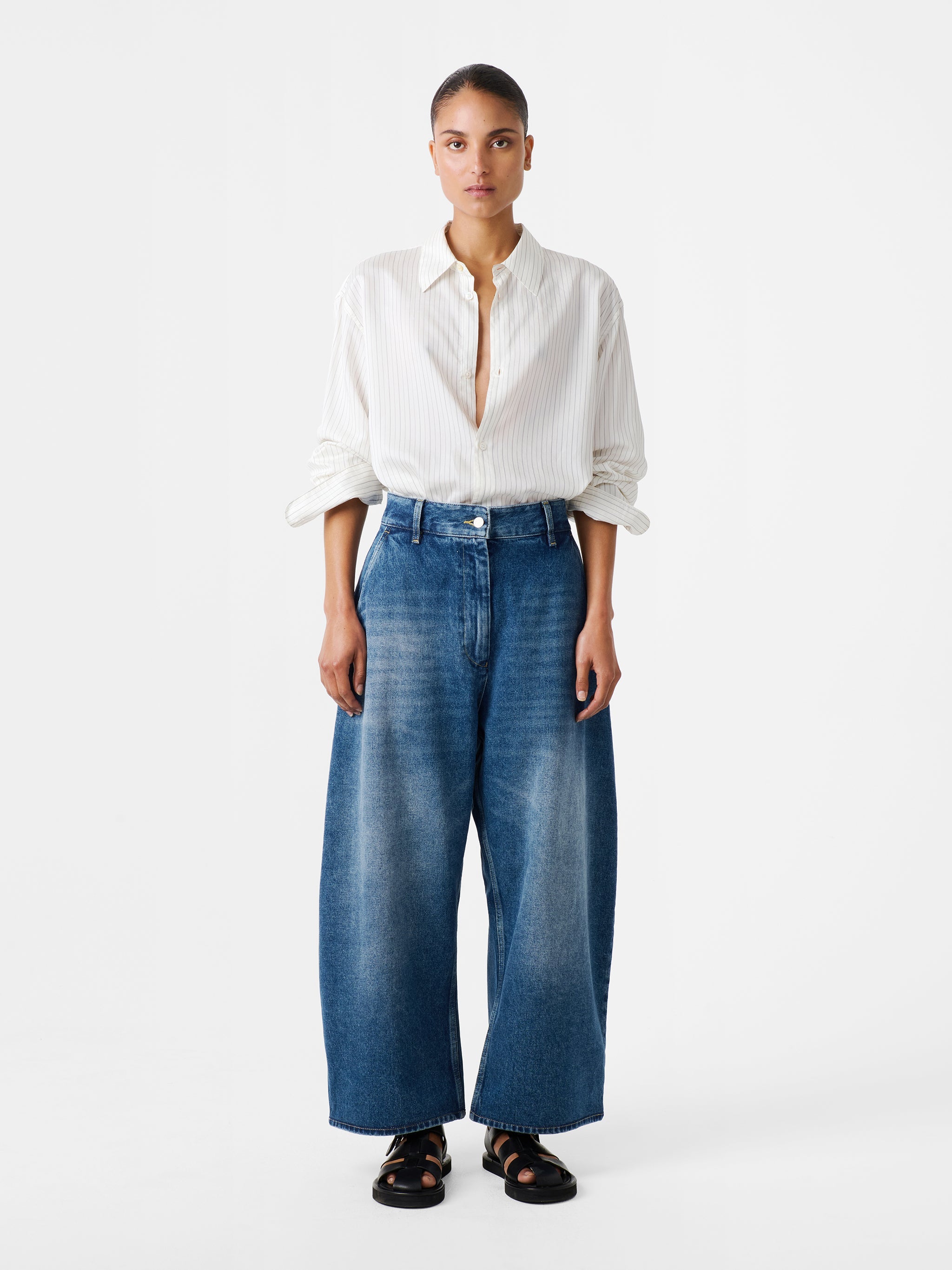 Woman standing wearing an elevated classic and wardrobe staple in the Studio Nicholson Worn Wash Indigo Chalco Wide Crop Denim Pant that are cut wide and cropped, with a curved leg and dropped crotch. They are constructed from a denim that’s been woven from regenerative cotton and made in the UK. 