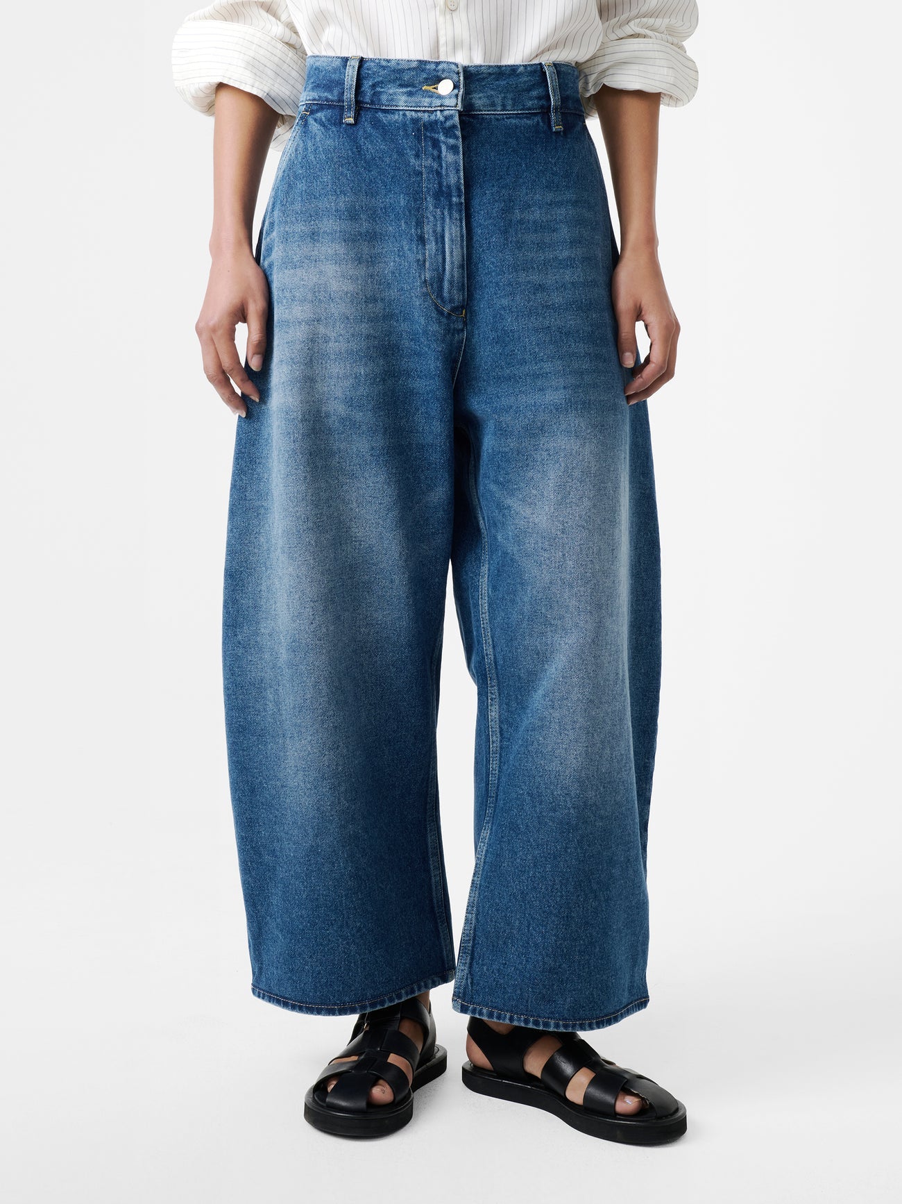 Close-up front view of a woman standing wearing an elevated classic and wardrobe staple in the Studio Nicholson Worn Wash Indigo Chalco Wide Crop Denim Jeans that are cut wide and cropped, with a curved leg and dropped crotch at Alas Eius. They are constructed from a denim that’s been woven from regenerative cotton and made in the UK.