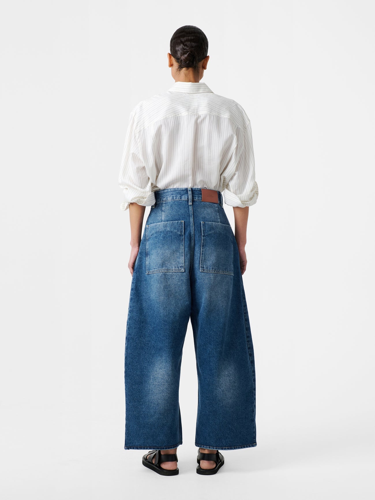Back view of a woman standing wearing an elevated classic and wardrobe staple in the Studio Nicholson Worn Wash Indigo Chalco Wide Crop Denim Pant that are cut wide and cropped, with a curved leg and dropped crotch at Alas Eius. They are constructed from a denim that’s been woven from regenerative cotton and made in the UK.