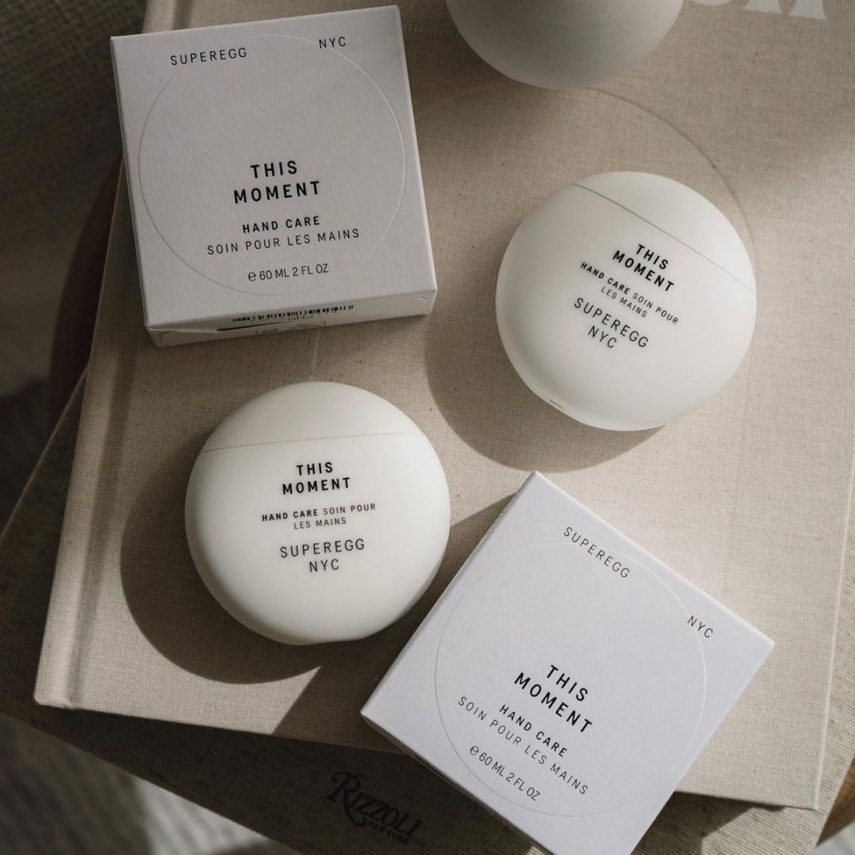 White boxes and containers of This Moment hand cream from the non-toxic skincare line called Sugeregg. The hand cream features a nature-aromatic treatment that conditions dry skin, targets discoloration, and replenishes elasticity for soft, silky hands at Alas Eius.