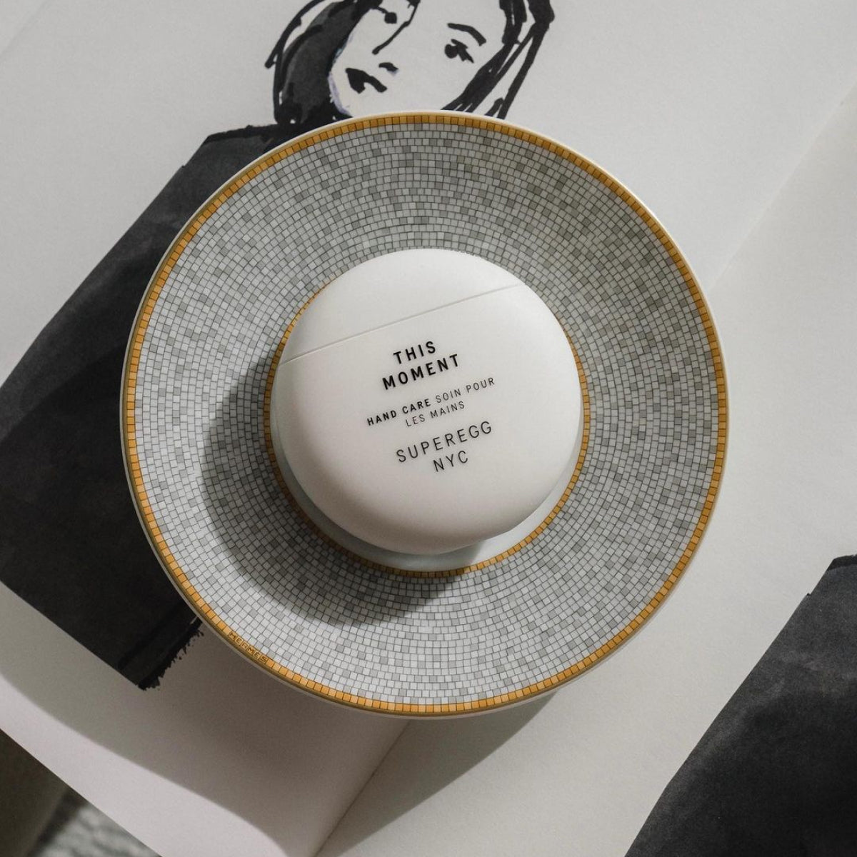 A round container of This Moment hand cream from the non-toxic skincare line called Sugeregg. The hand cream features a nature-aromatic treatment that conditions dry skin, targets discoloration, and replenishes elasticity for soft, silky hands at Alas Eius.