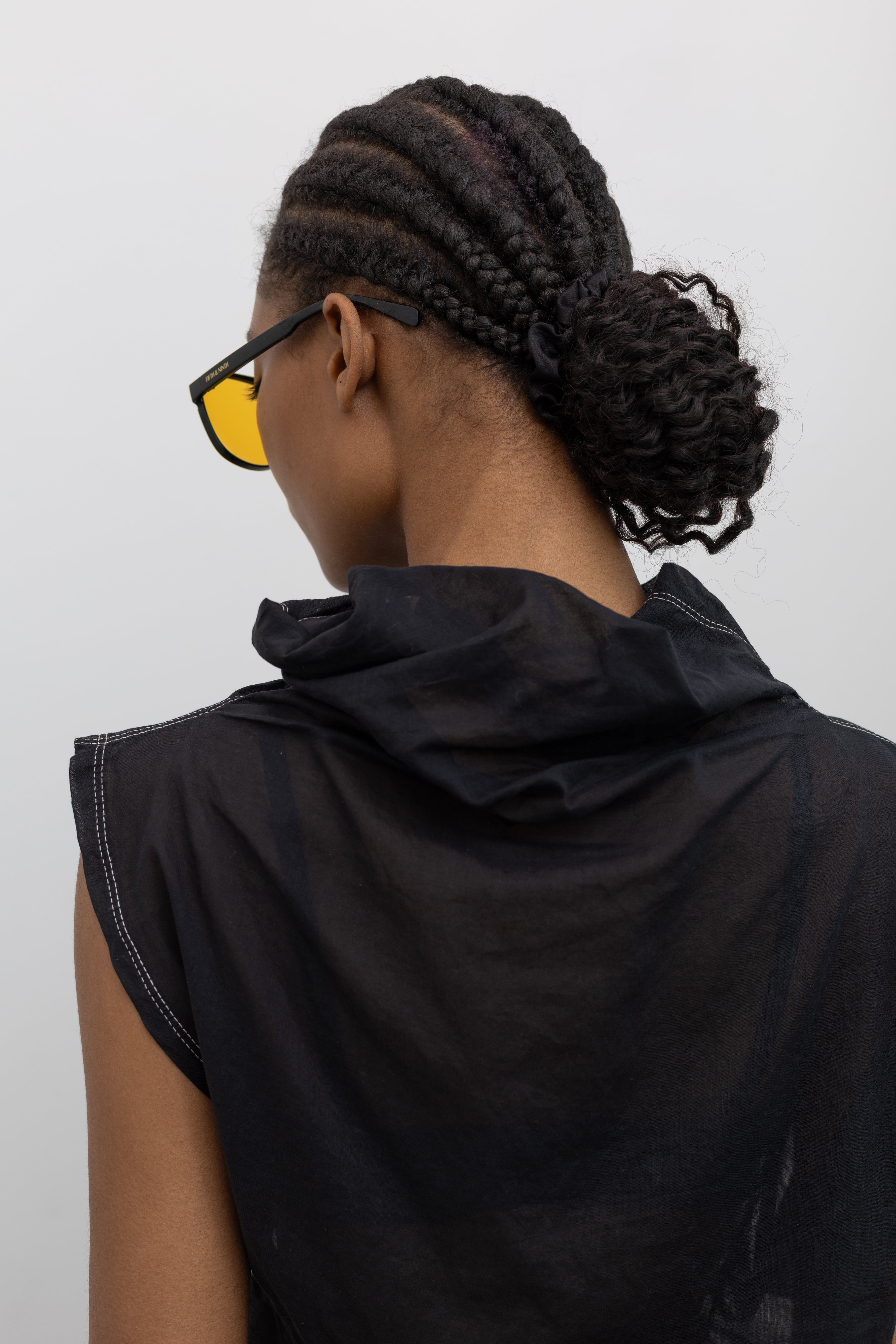 Close-up back view of a woman posing wearing a unique Tela brand semi-sheer lightweight washed muslin black top featuring a soft draped asymmetrical neckline paired with a classic wide black leather belt on black wide leg trousers at an Alas Eius store. 
