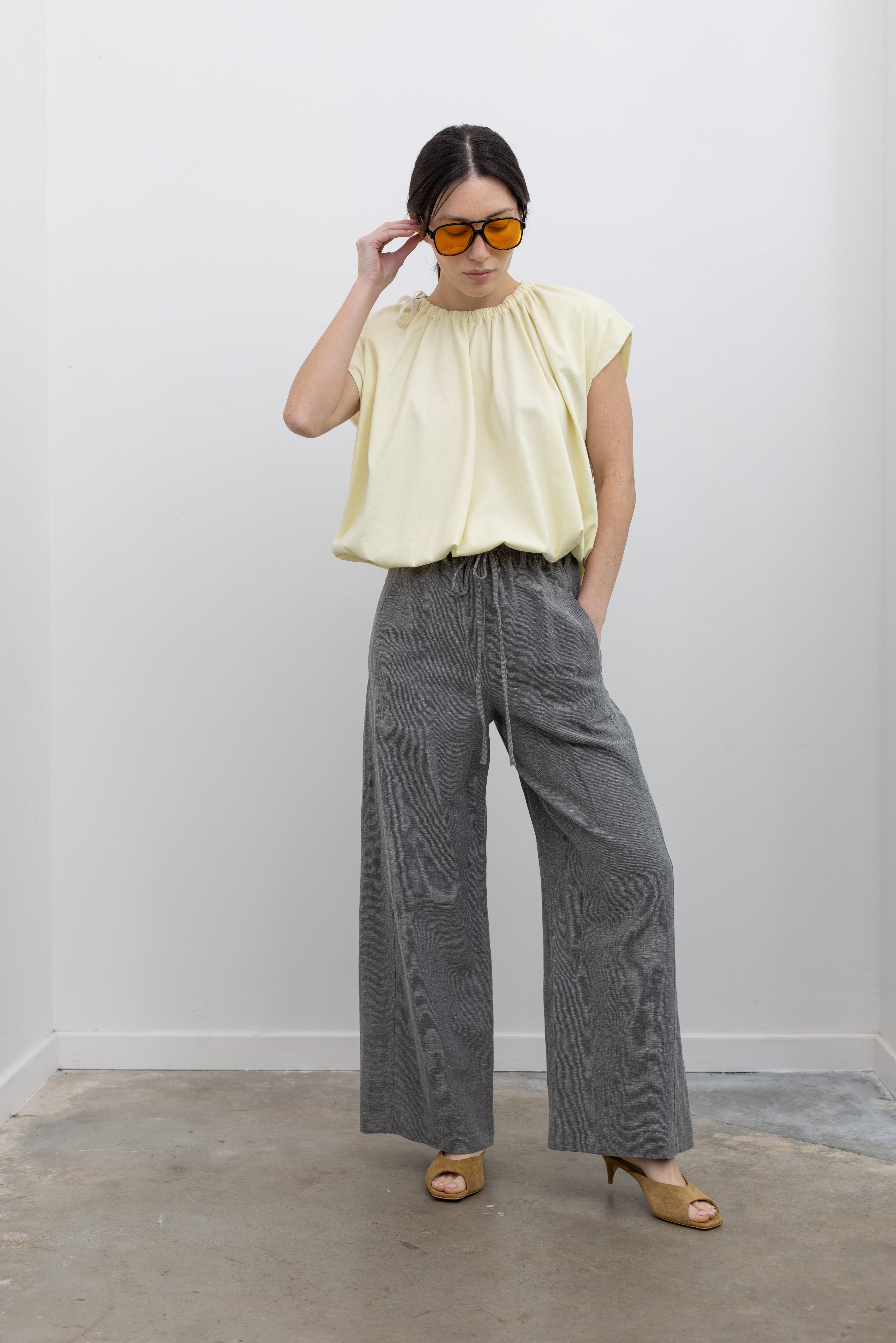 Front view of a woman posing with sunglasses wearing sophisticated spring fashion in a Tela Light Yellow Gathered Neck Bubble Hem Top, Tela Lucio light gray drawstring pants featuring a wide leg and side seam pockets, and Tela Feng Mustard Leather Heeled Sandals at an Alas Eius store. 