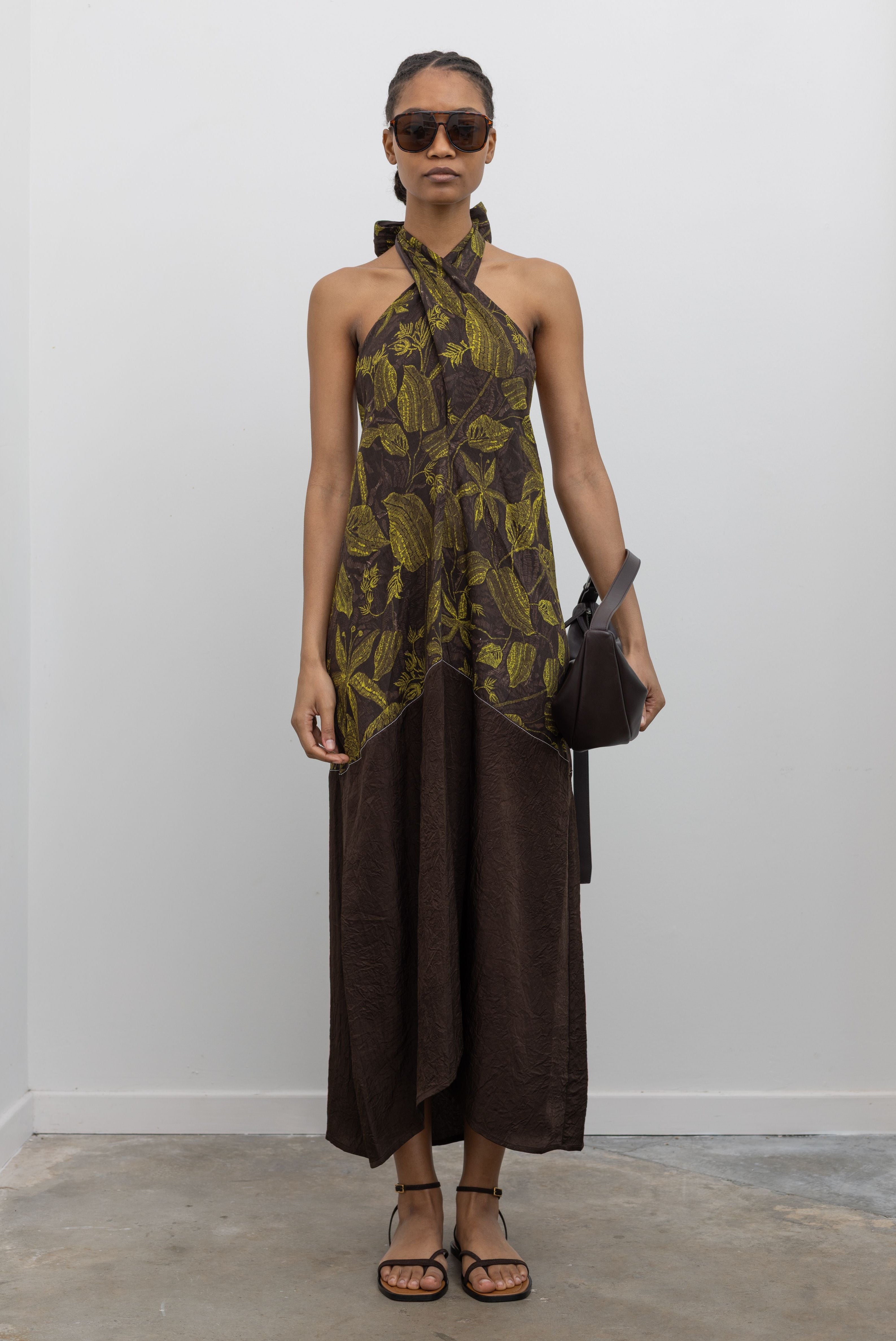 Full length front view of a woman wearing the perfect summer vacation dress in a Tela label olive green botanical print halter neck midi dress with a contrasting chocolate brown hem paired with ATP strappy leather sandals and a leather St. Agni bag at an Alas Eius Store.