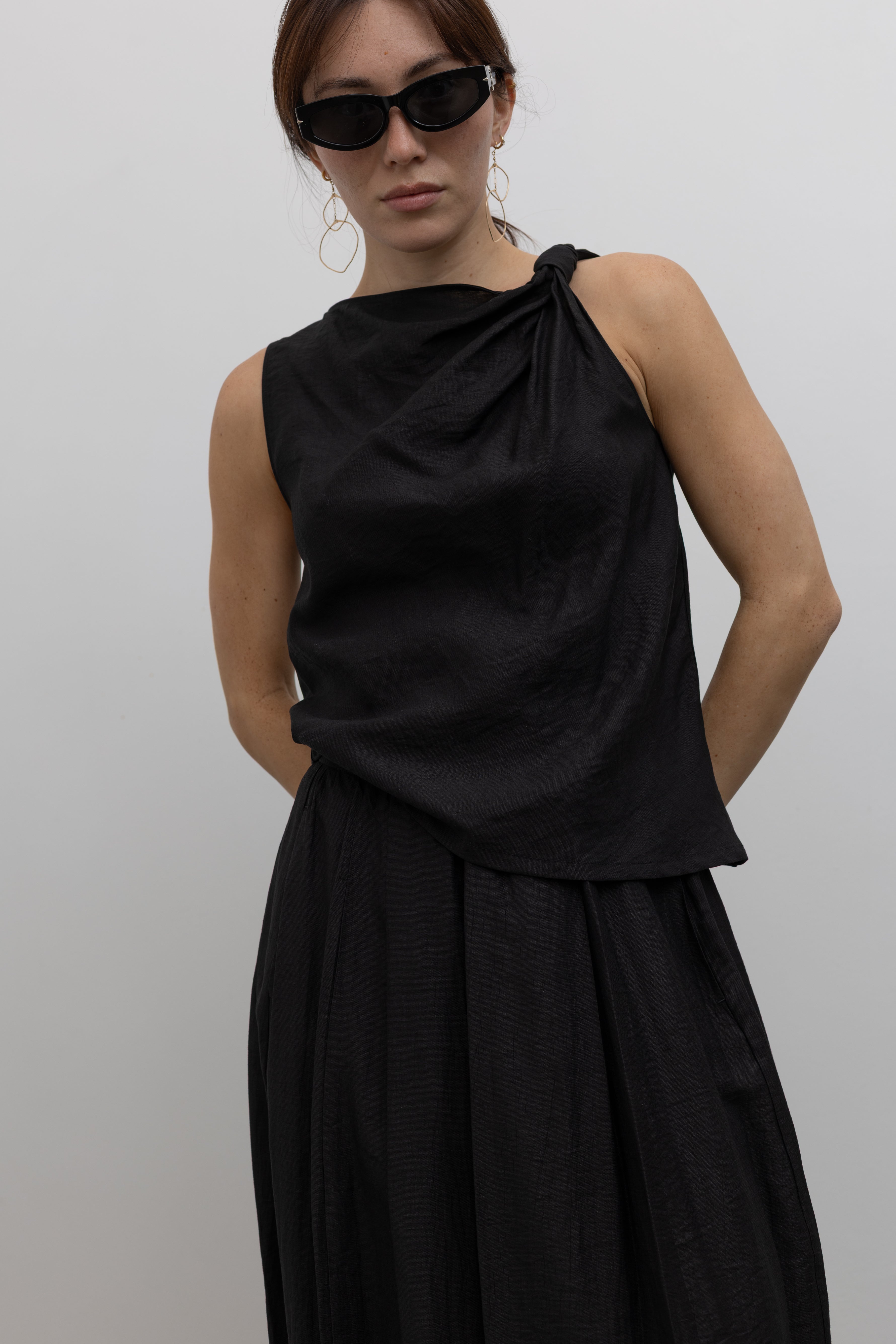 Woman standing wearing Spring fashion in an elegant and flattering Tela Cody black linen knotted shoulder sleeveless top at Alas Eius. 