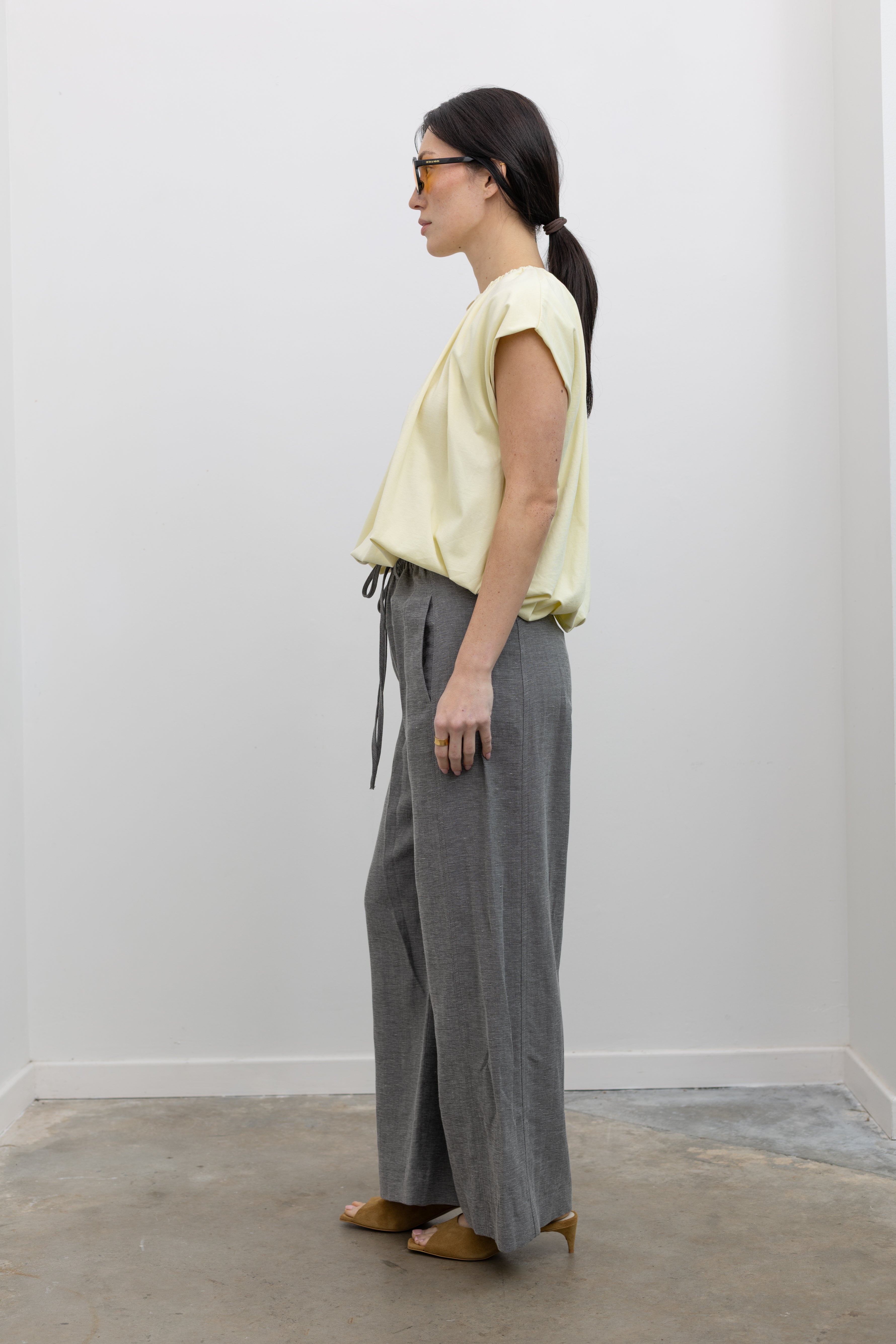 Full length side view of a woman standing wearing modern minimalist spring fashion in a Tela Light Yellow Gathered Neck Bubble Hem Top, Tela Lucio light gray drawstring pants featuring a wide leg and side seam pockets, and Tela Feng Mustard Leather Heeled Sandals at an Alas Eius store. 
