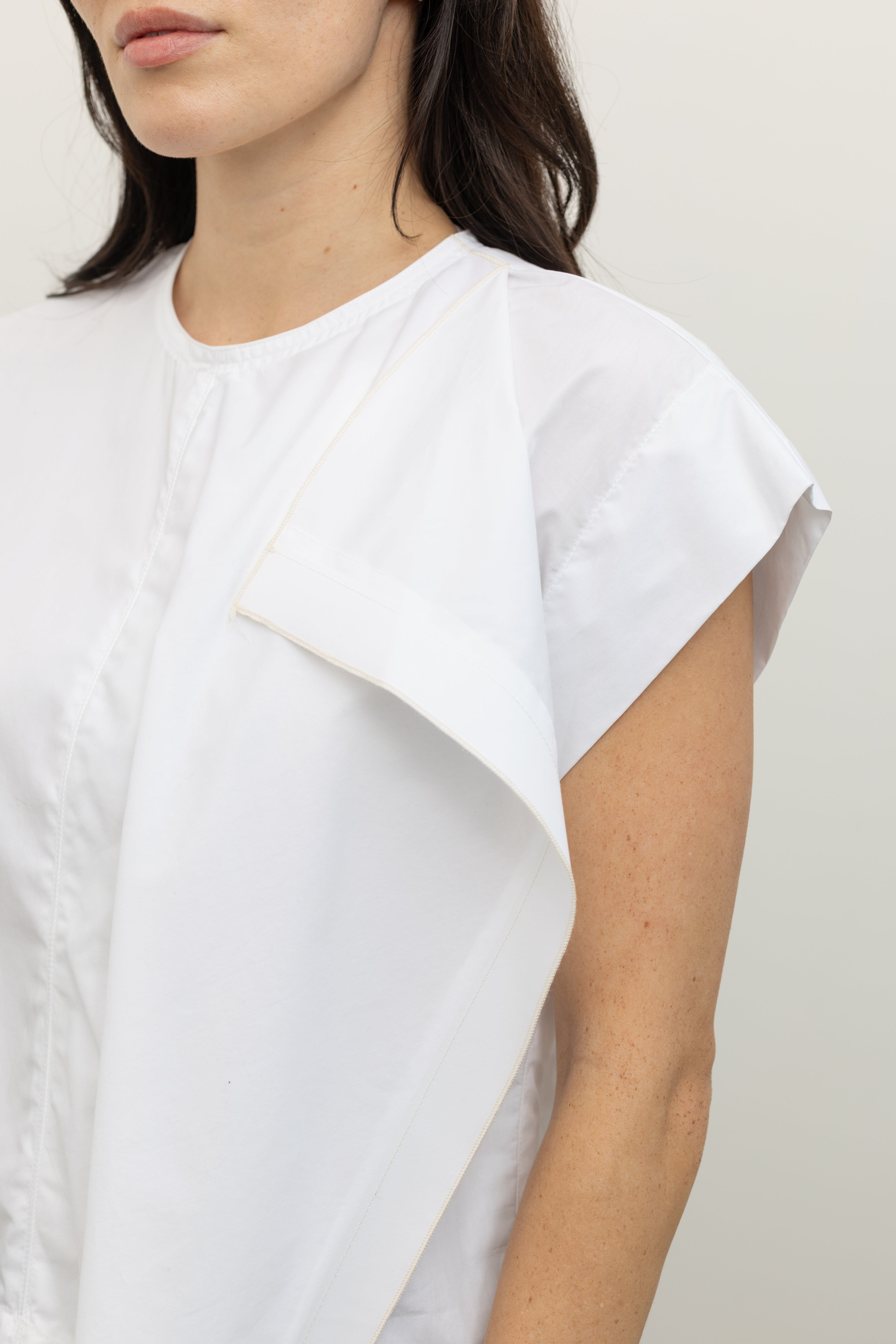 Close-up product view of a woman wearing contemporary spring fashion in a Tela brand white Livia shirt featuring a front pleat that creates an intentional asymmetrical drape, crew neckline and relaxed cap sleeves paired with oversized baggy barrel leg pants at at an Alas Eius store. 