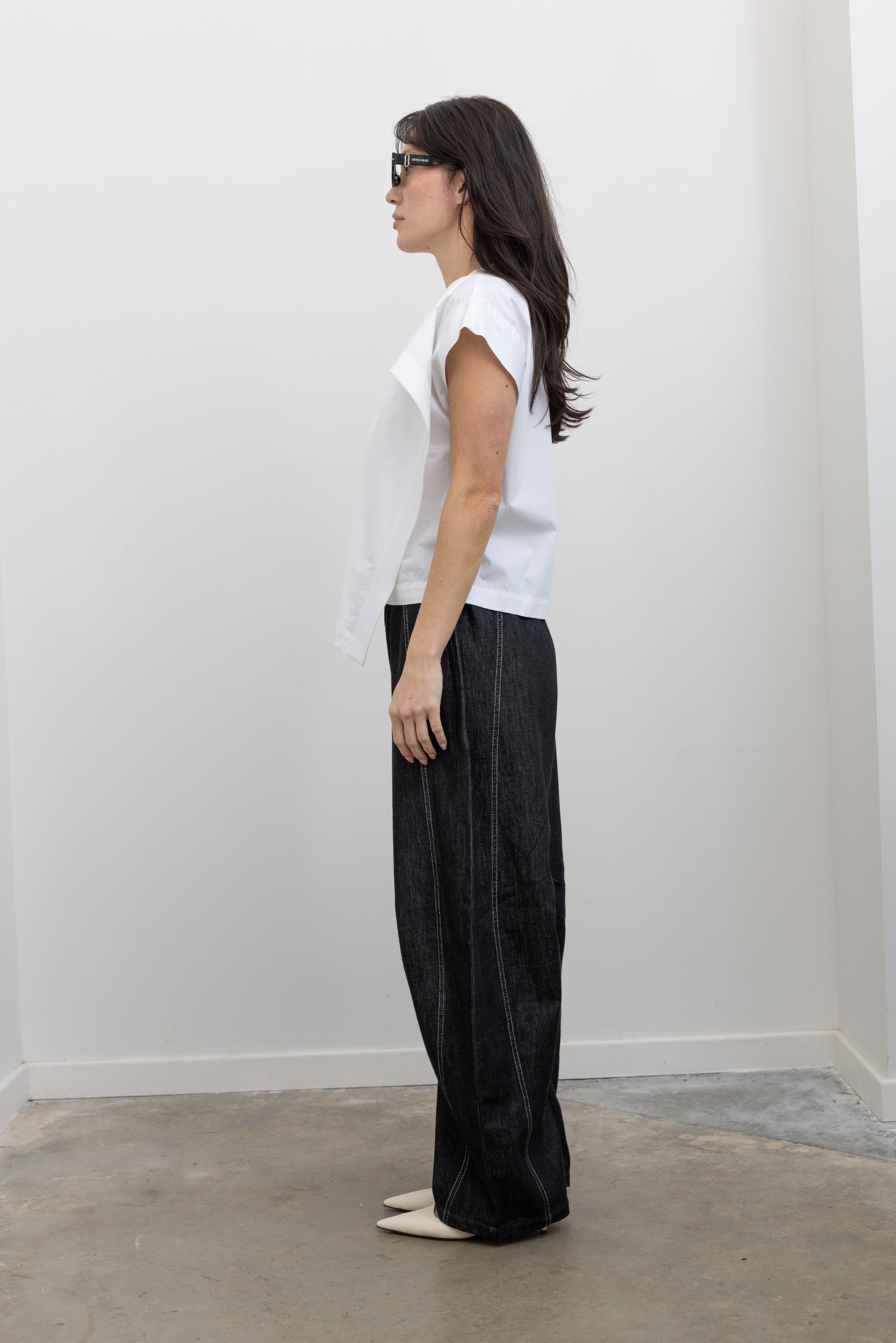 Full length side view of a woman wearing modern minimalist spring fashion in a Tela brand white Livia shirt featuring a front pleat that creates an intentional asymmetrical drape, crew neckline and relaxed cap sleeves paired with oversized baggy barrel leg pants at at an Alas Eius store. 