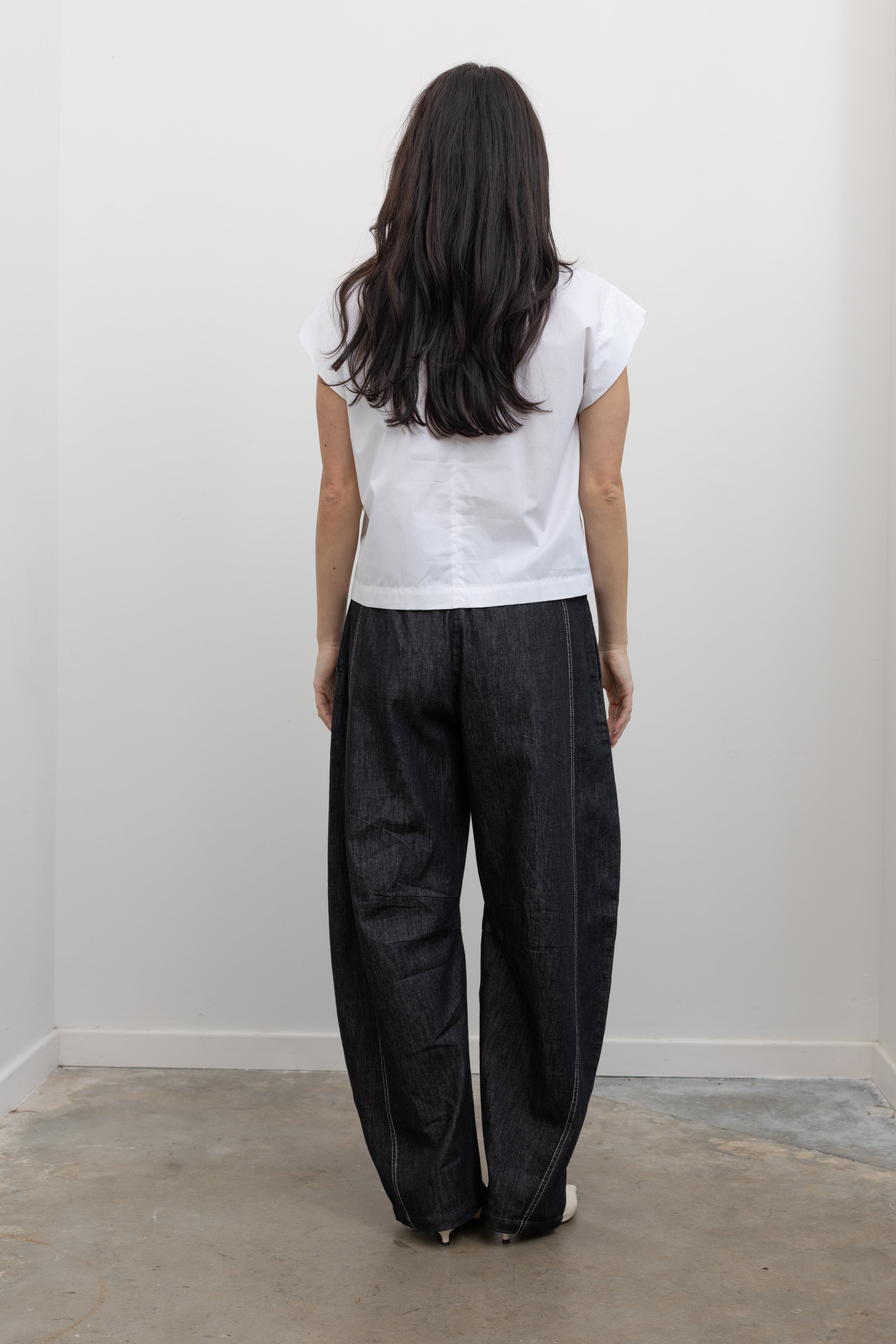 Full length back view of a woman wearing modern minimalist spring fashion in a Tela brand white Livia shirt featuring a front pleat that creates an intentional asymmetrical drape, crew neckline and relaxed cap sleeves paired with oversized baggy barrel leg pants at at an Alas Eius store. 