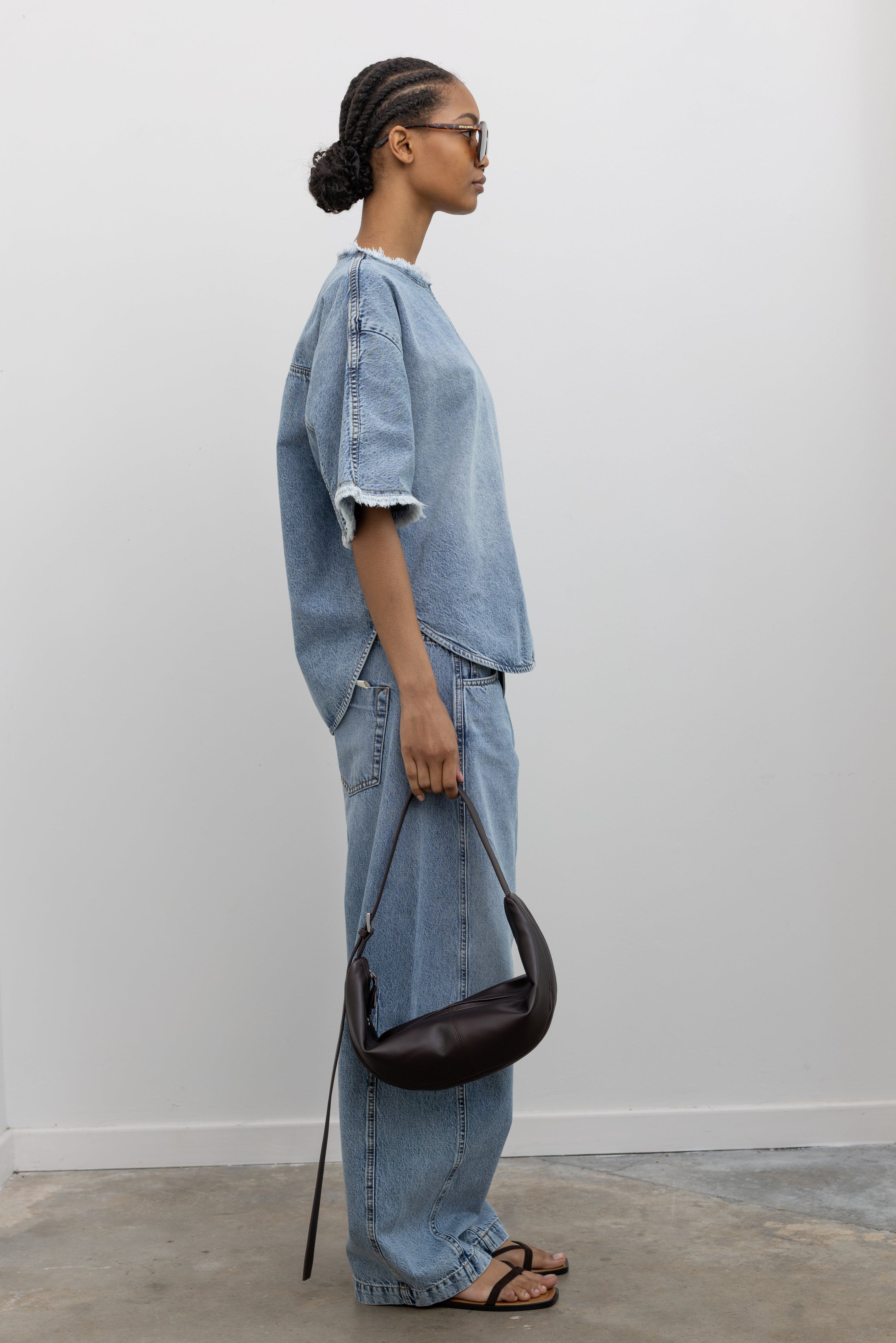 Full length side view of a Woman wearing modern spring streetwear fashion in a Tela Light Blue Denim Oversized Short Sleeve Shirt featuring  a clean light-blue wash, distinct raw-edge detailing, and a minimalist back neck closure paired with light-blue wash denim jeans and a leather St. Agni bag at an Alas Eius store. 