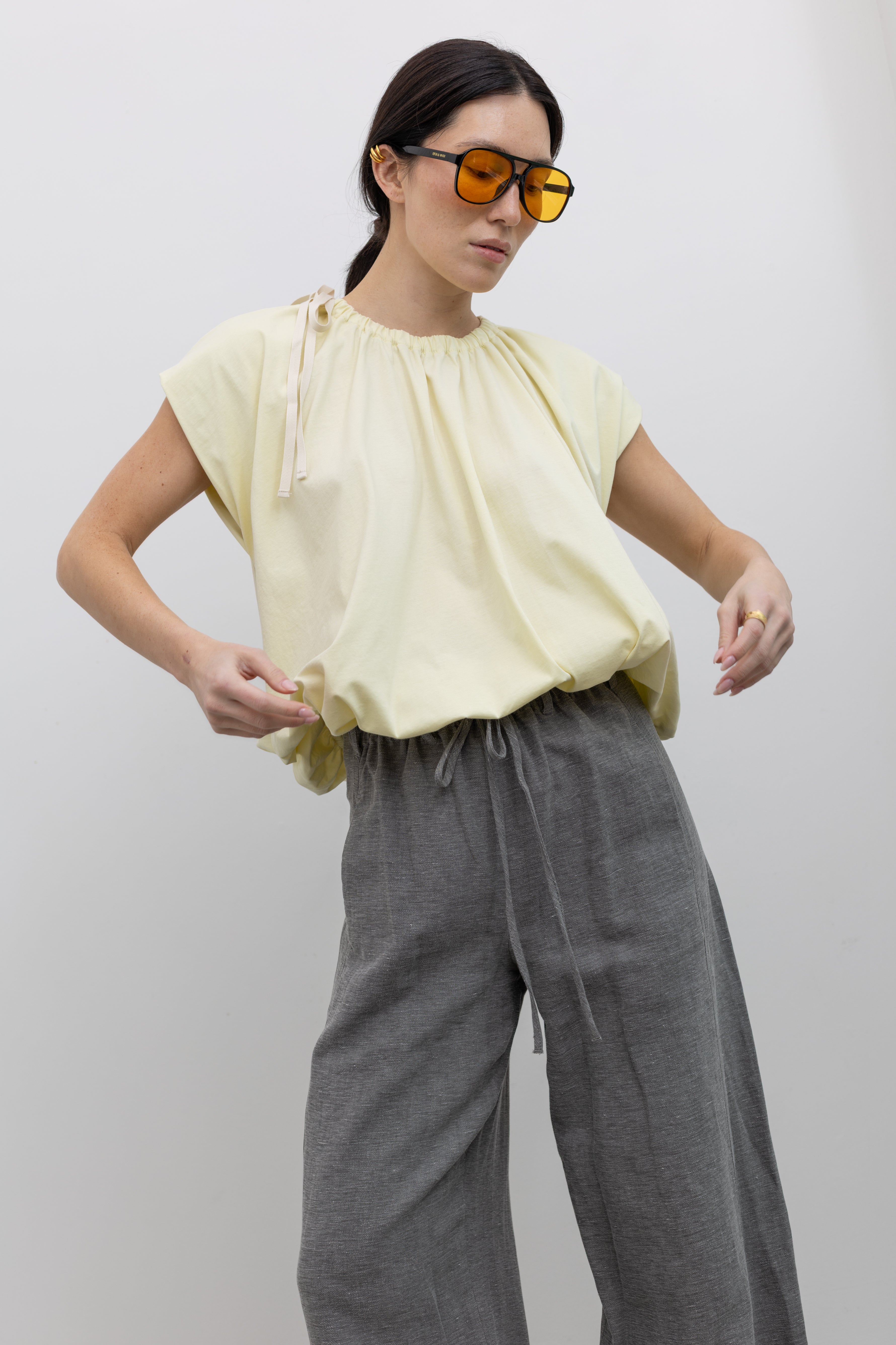 Front view of a woman in motion wearing contemporary spring fashion in a Tela Light Yellow Gathered Neck Bubble Hem Top, Tela Lucio light gray drawstring pants featuring a wide leg and side seam pockets, and Tela Feng Mustard Leather Heeled Sandals at an Alas Eius store. 